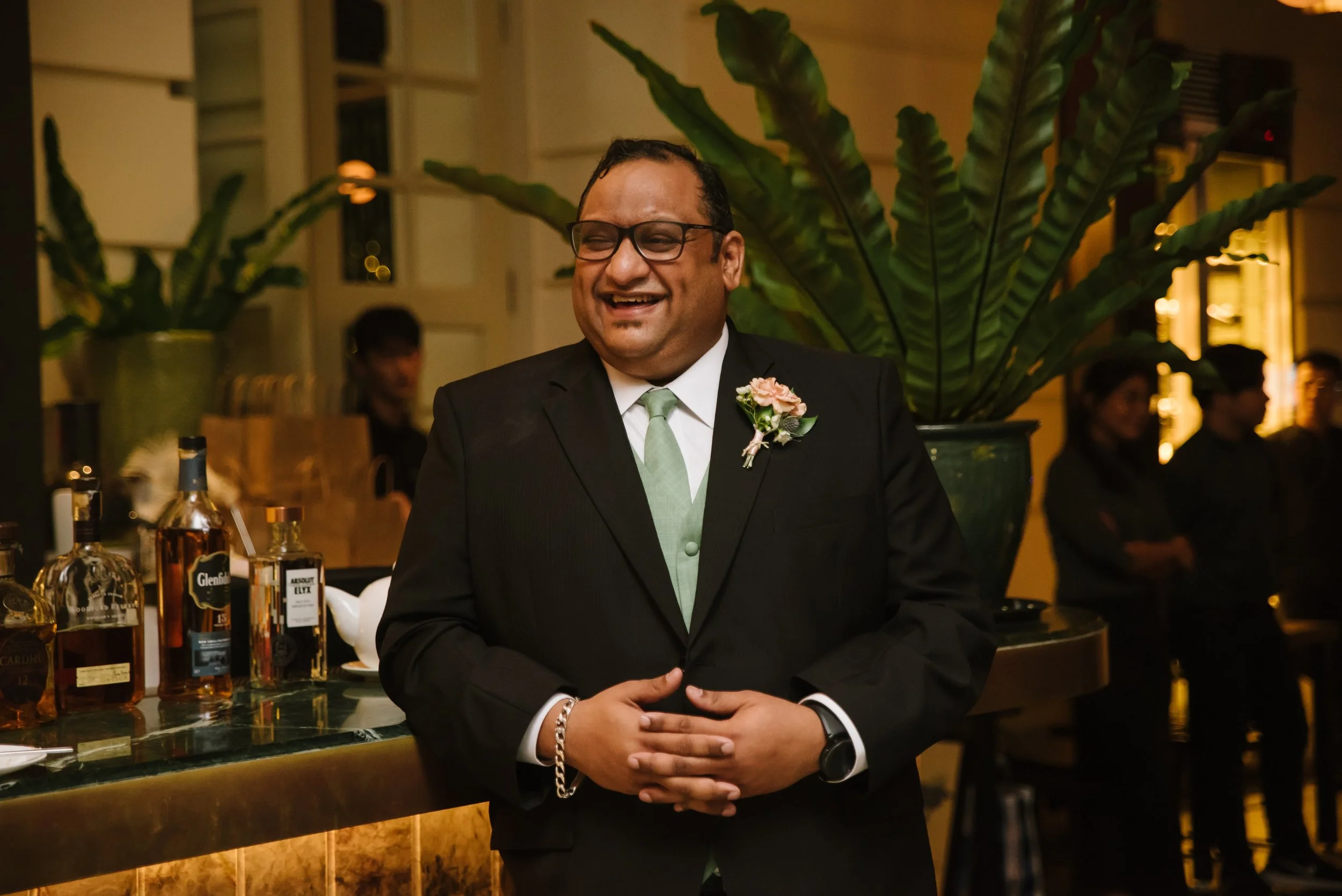 A man in a black suit with a light green tie and glasses, smiling, standing next to a bar with various liquor bottles, in a warmly lit room with large green plants and other people in the background.