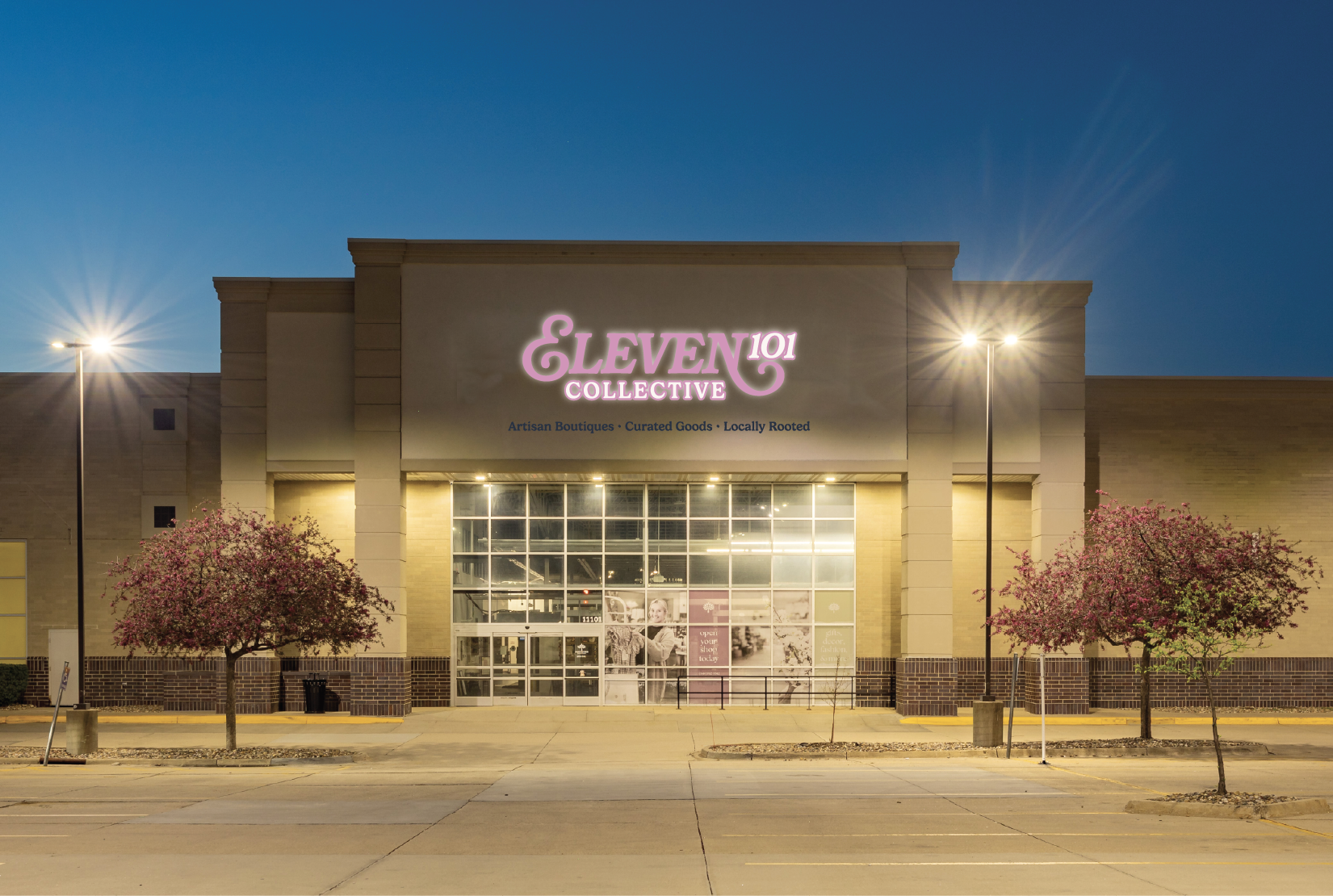 The storefront of Eleven 101 Collective, a retail store, is illuminated at nighttime with a bright pink and white sign. The store features large glass windows and a small parking lot with two blooming purple trees on either side of the entrance.