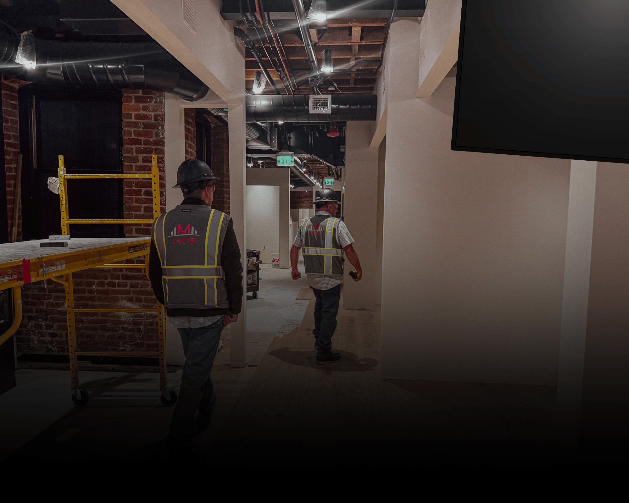 Two construction workers wearing safety helmets and vests working inside a building under renovation. The background shows exposed brick walls, ductwork, and unfinished ceiling with pipes and wires.