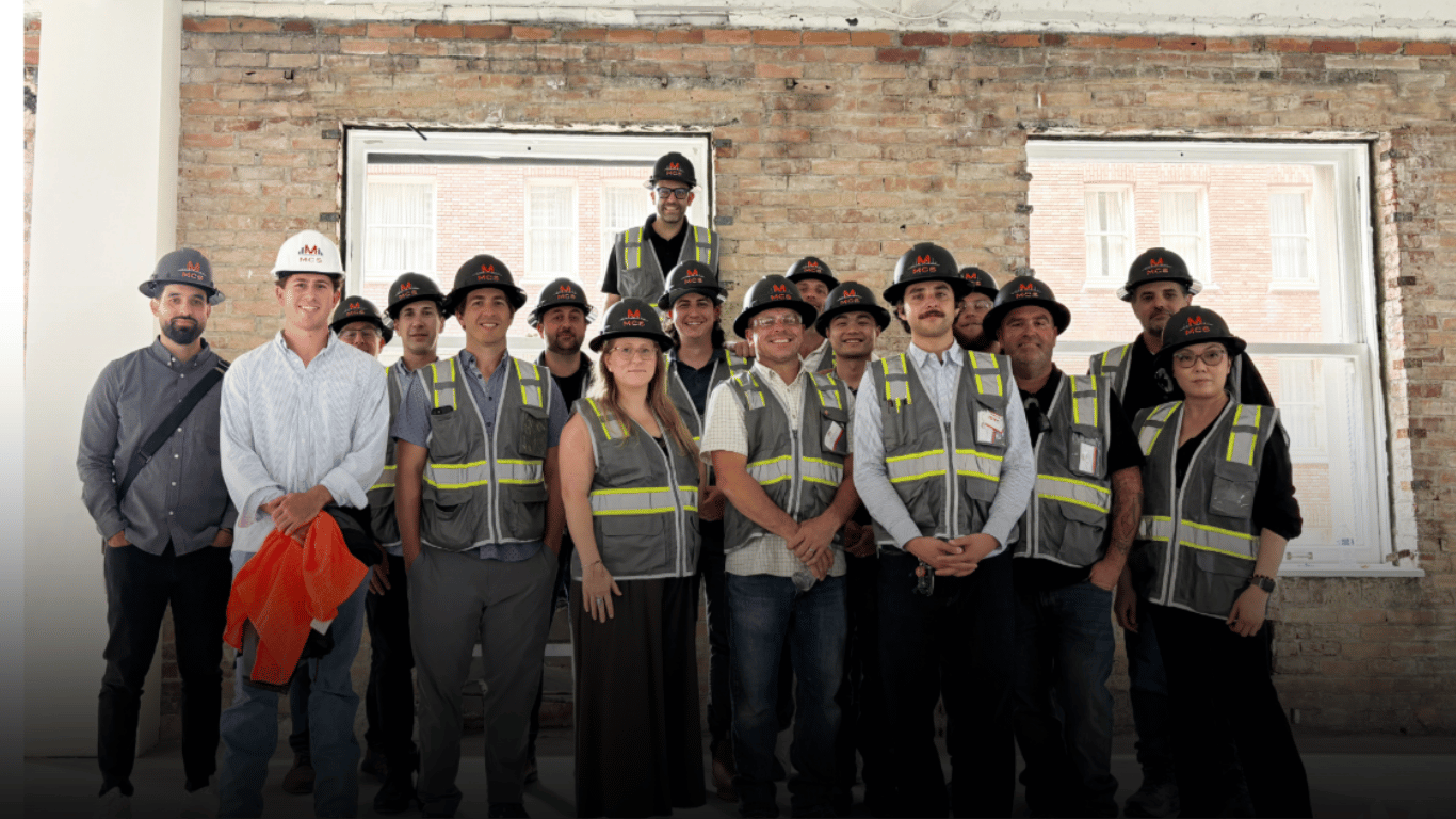 MCS construction team in safety vests and helmets inside a partially finished building with brick walls and large windows.