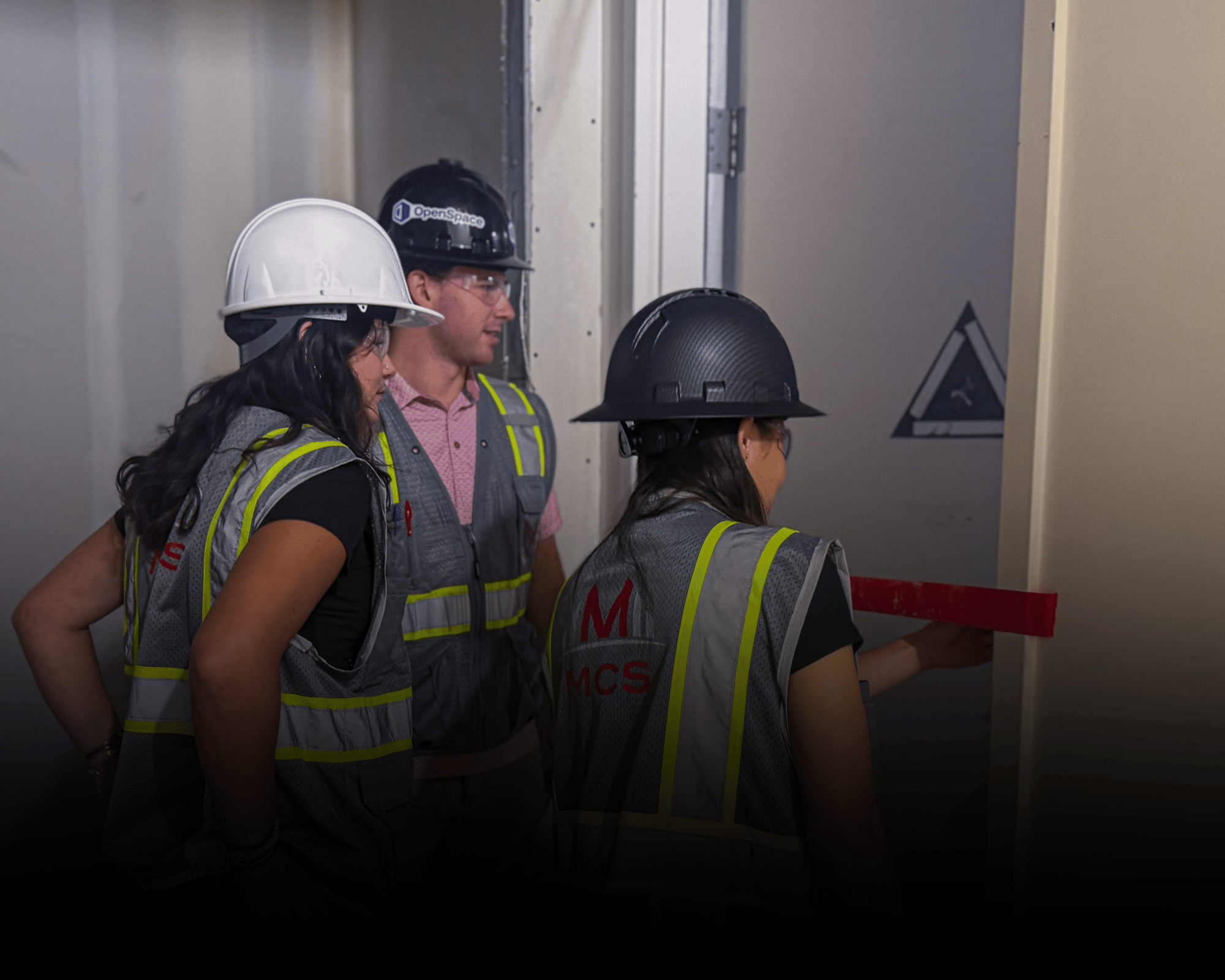 Four MCS construction workers wearing safety gear, hard hats, and reflective vests, inspecting an elevator door in a building under construction.