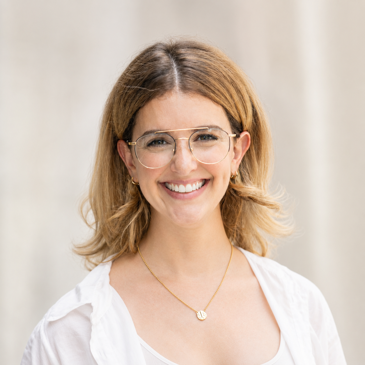 A smiling woman with shoulder-length blonde hair, wearing round glasses, gold jewelry, and a white top, standing against a neutral background.