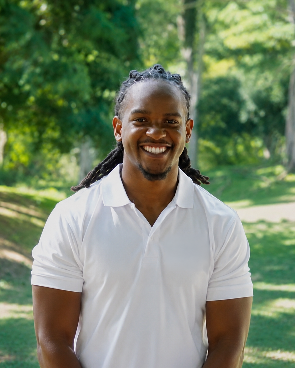 A smiling man with long dreadlocks in a white polo shirt standing outdoors in a park with green trees in the background.