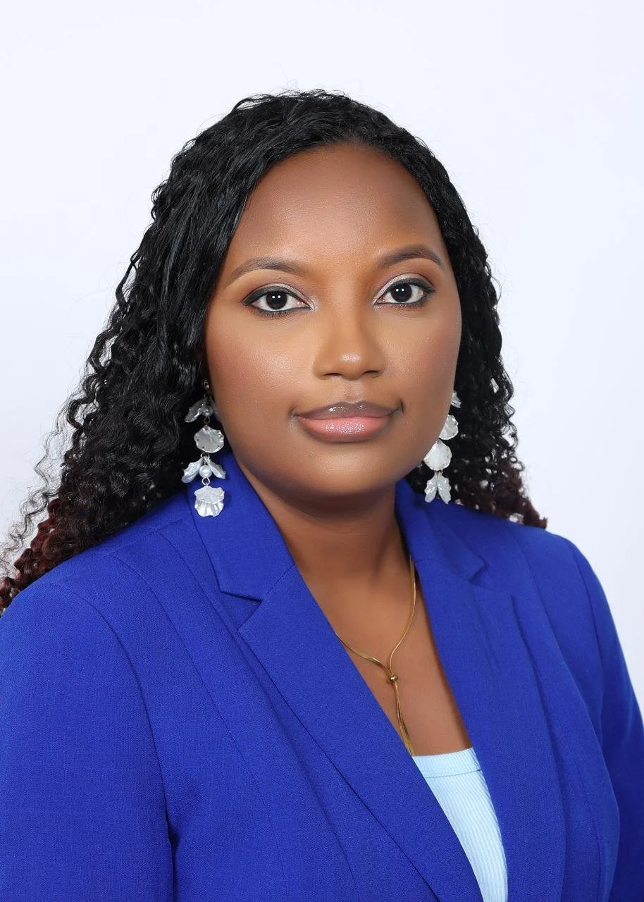 A professional woman with dark curly hair, wearing a blue blazer, white top, silver earrings, and a gold necklace, posing against a light background.