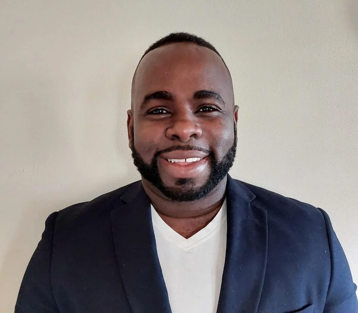 A man with a short beard smiling for a photo, wearing a navy blazer over a white shirt, standing against a plain light-colored wall.