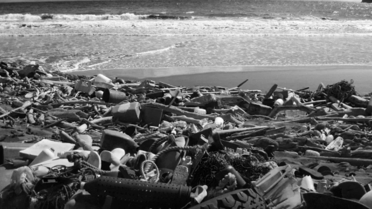 Beach with scattered trash and debris, ocean waves in the background, black and white photo.