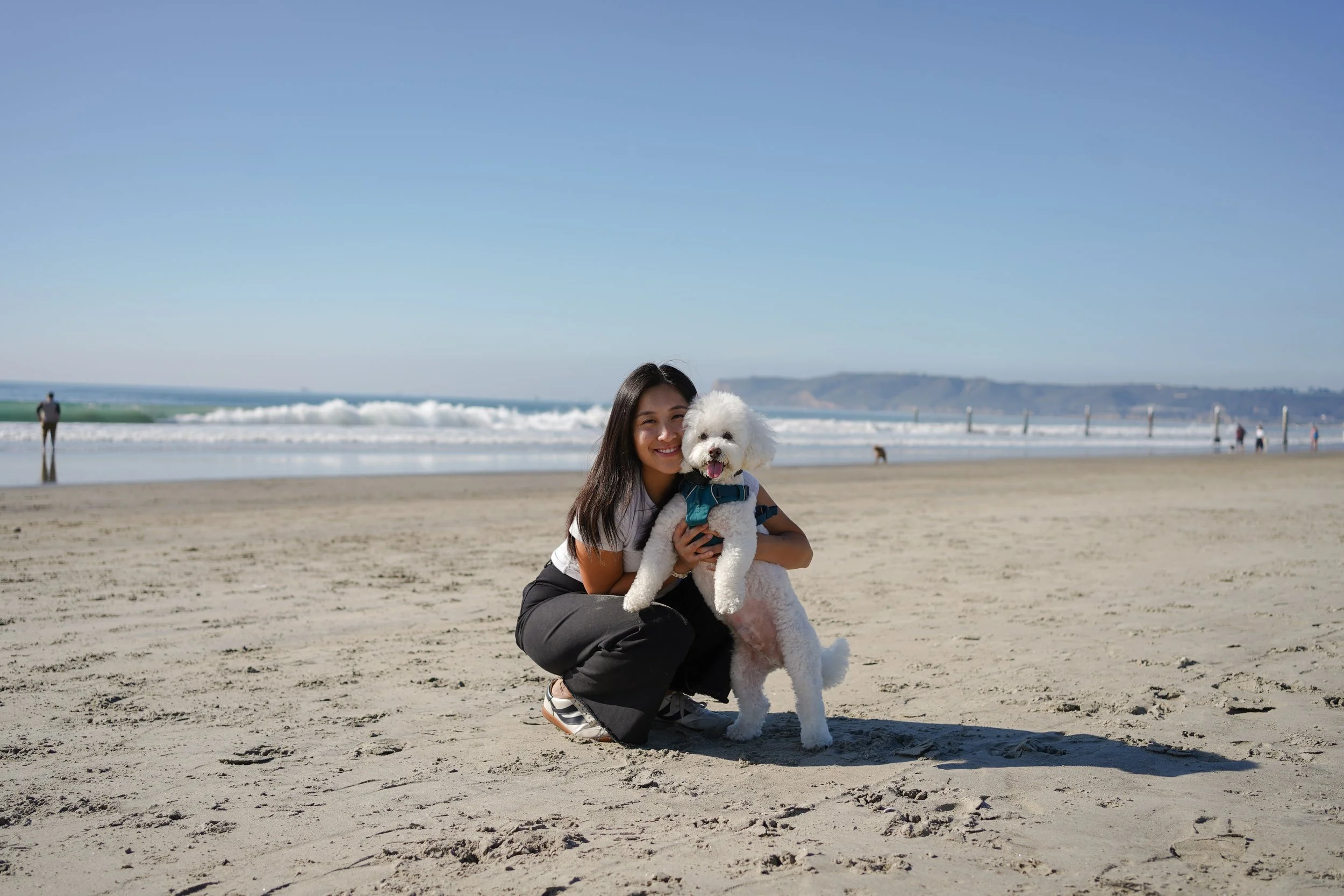 A woman kneeling on the sandy beach, smiling and holding a white fluffy dog wearing a blue harness. The ocean and distant hills are visible in the background, with a few people walking along the shoreline.