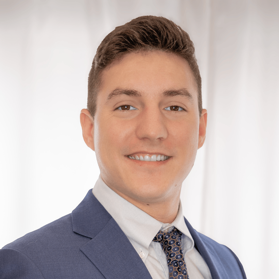 Professional portrait of a young man with short brown hair, wearing a navy suit, white shirt, and patterned tie, smiling against a blurred light background.