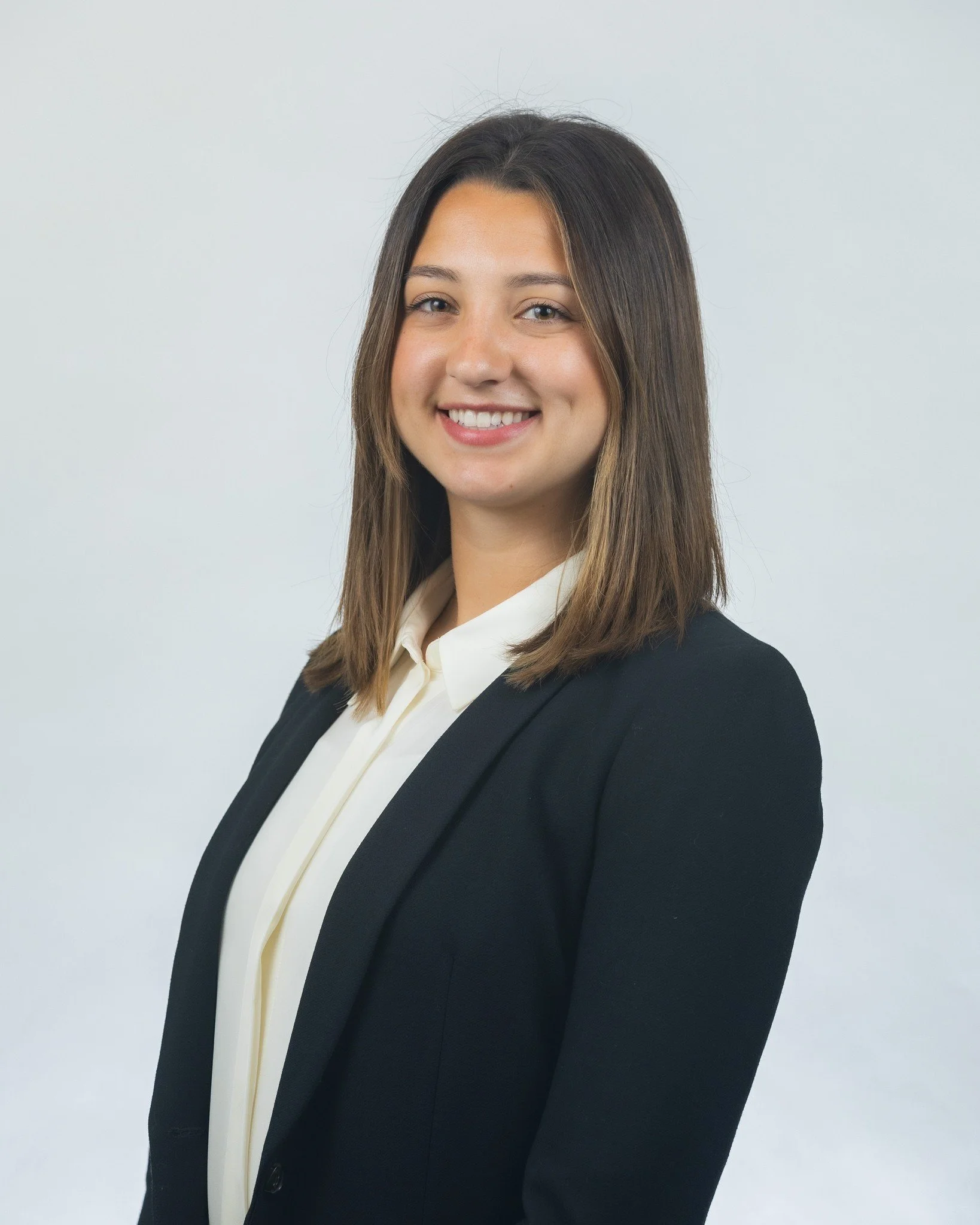 A young woman with straight brown hair wearing a black blazer, white blouse, and standing against a plain light grey background, smiling at the camera.