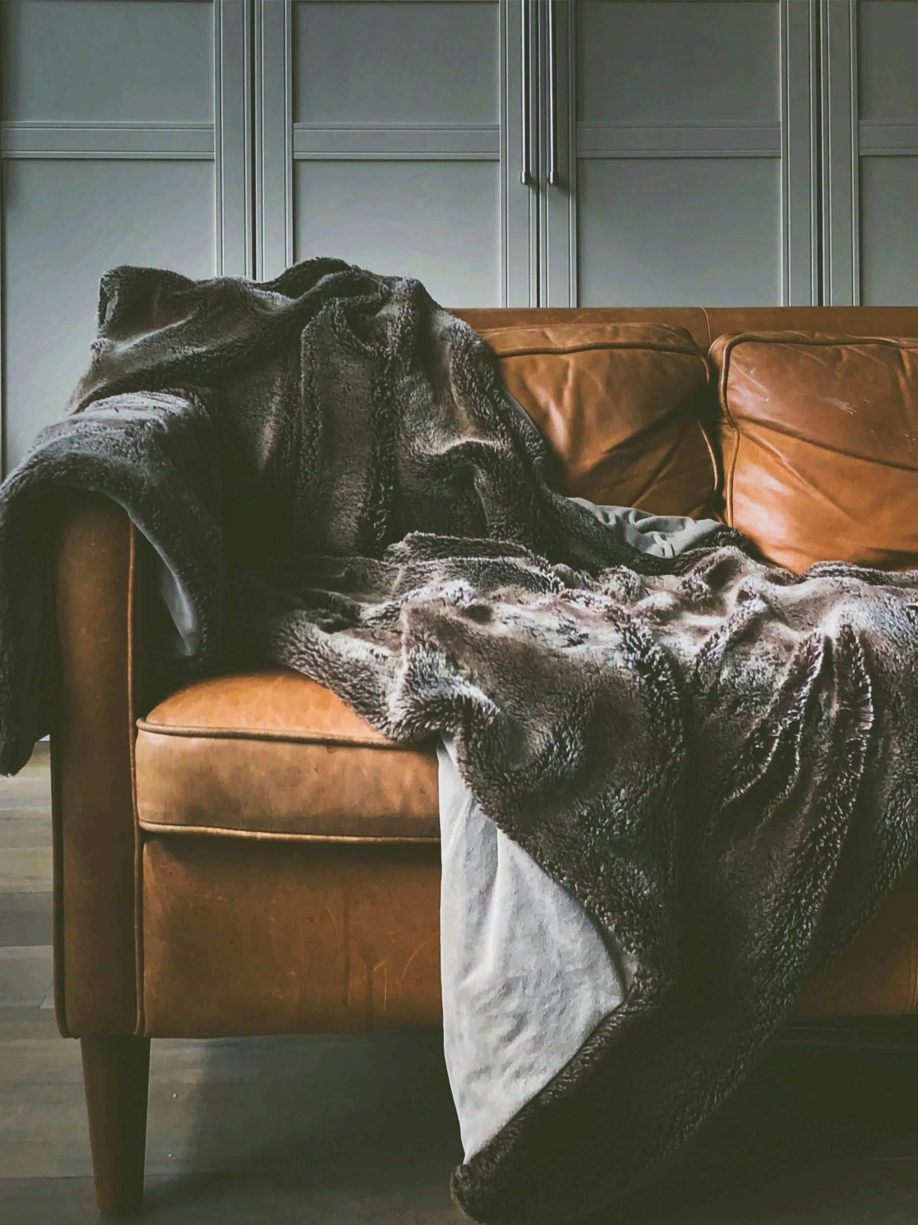 Close-up of a brown leather sofa with a black and gray patterned blanket draped over the armrest, against a background of gray cabinets.