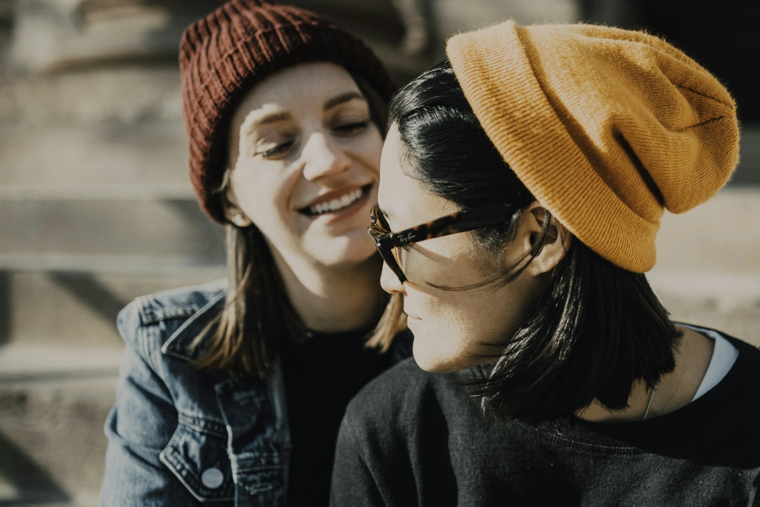 Two women, one with a red beanie and another with an orange beanie, face each other closely and smile outdoors.