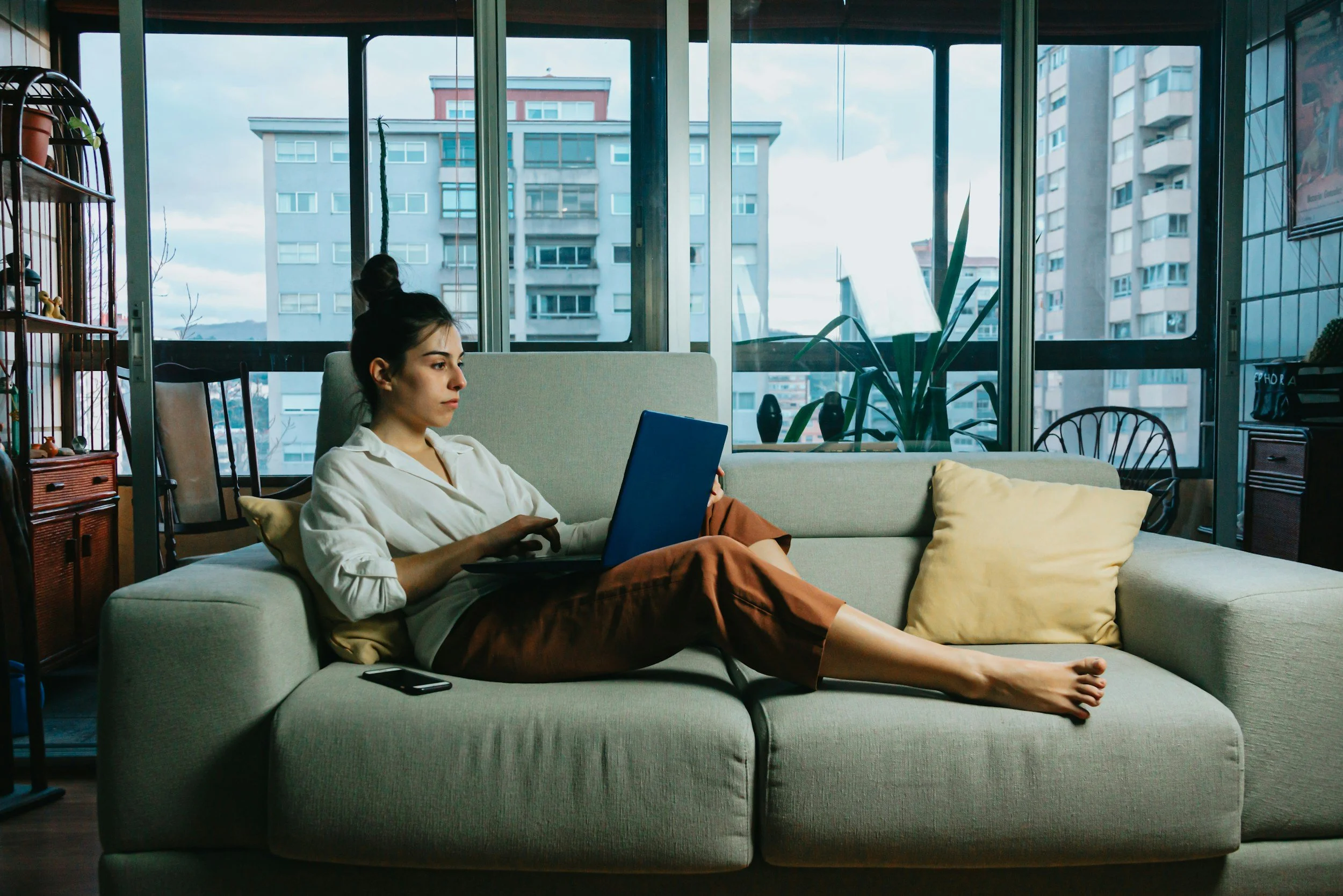 Young woman sitting on a sofa in a sunlit apartment, using a laptop, with a large window showing city buildings in the background.