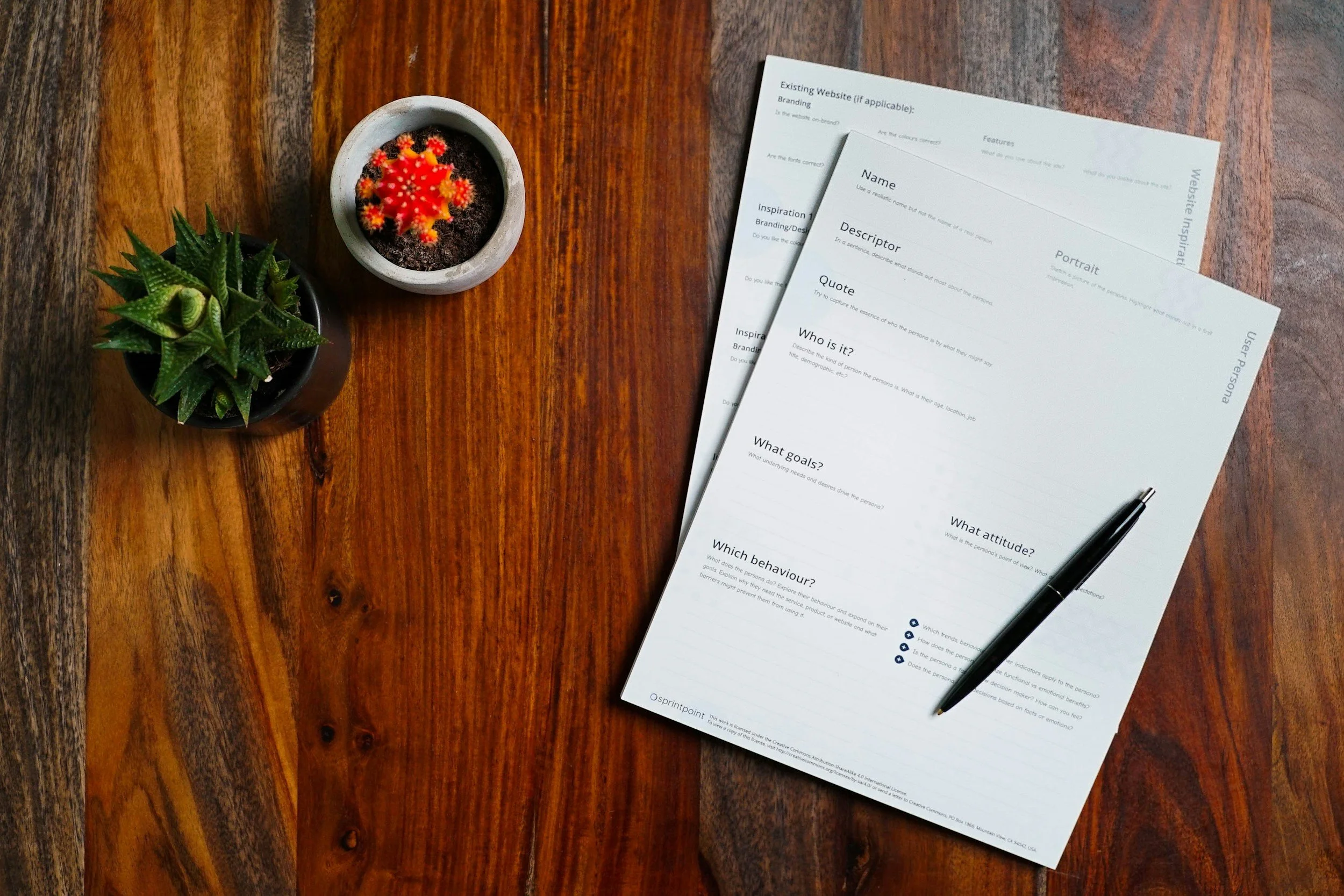 A wooden table with two small potted cacti and several sheets of paper with questions, along with a black pen.