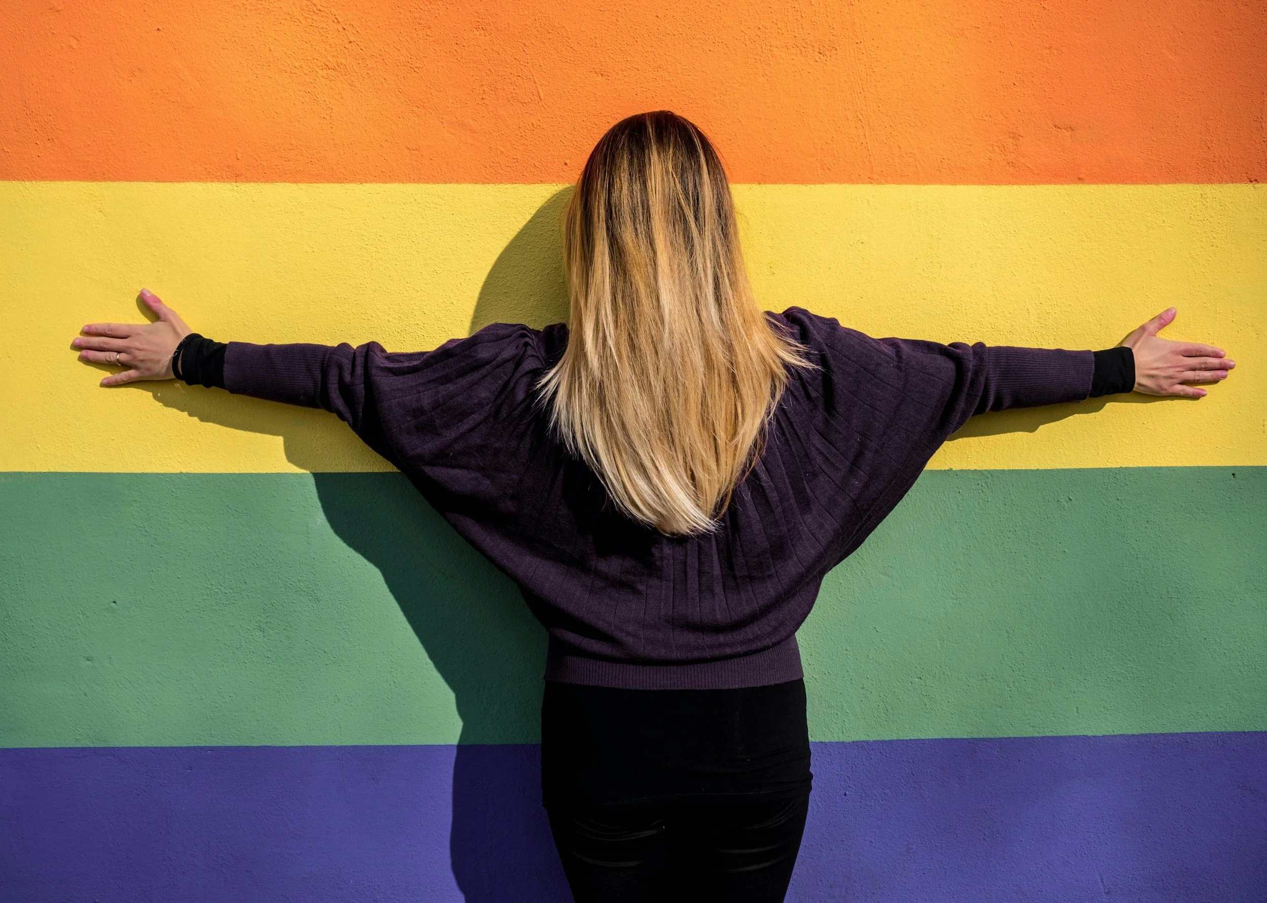 Person with long blonde hair wearing a dark hoodie, standing with arms outstretched, facing a rainbow-colored wall.