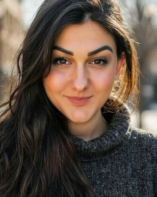 Close-up of a young woman with long dark hair, smiling slightly while looking at the camera outdoors.
