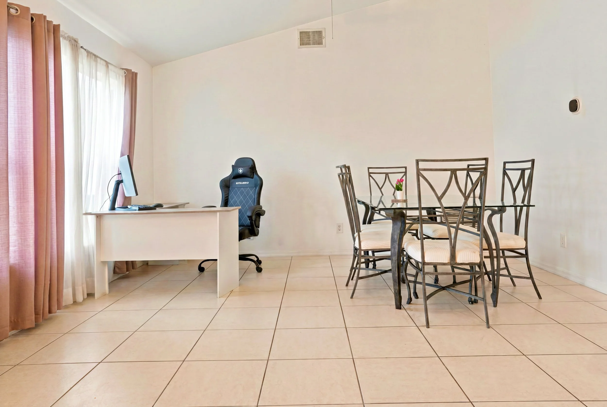 A room with beige tiled floor, white walls, and curtains on windows. There is a white desk with a computer monitor, keyboard, and mouse, and a black gaming chair. Adjacent to the desk is a glass-top dining table with six metal chairs, one with a smal