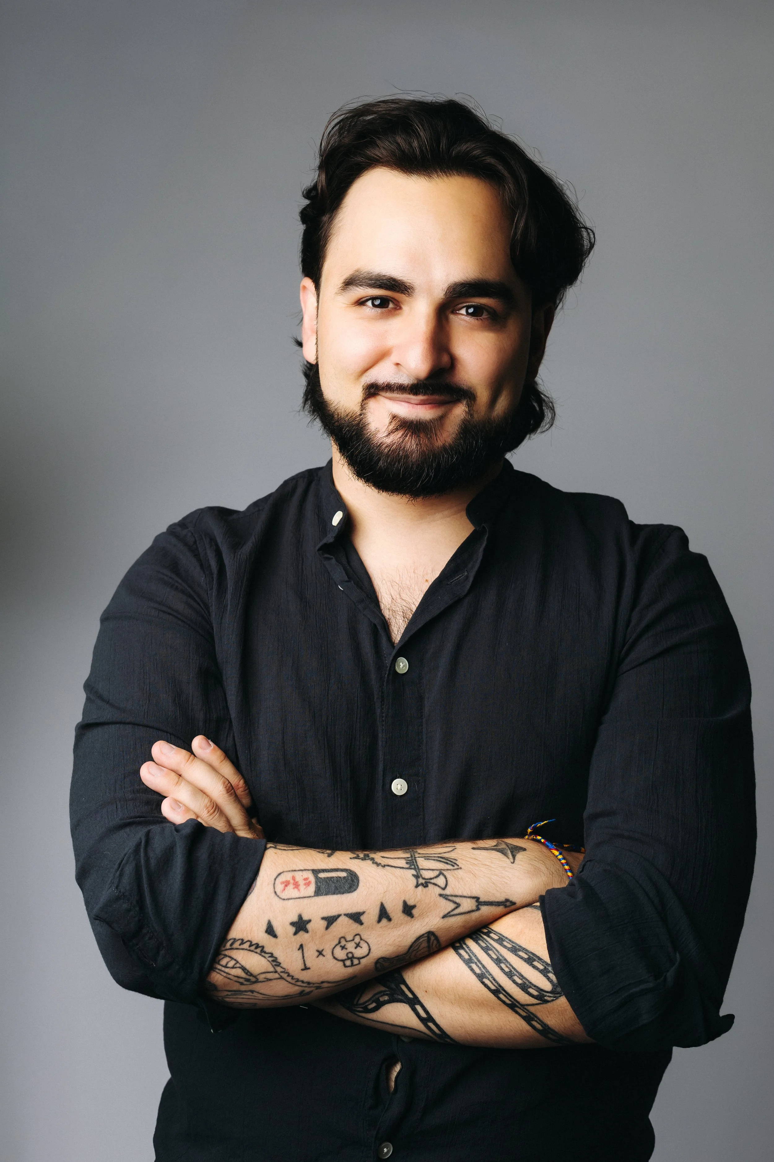 A man with dark hair, a beard, and tattoos on his arms, wearing a black shirt, standing with arms crossed against a gray background, smiling slightly.