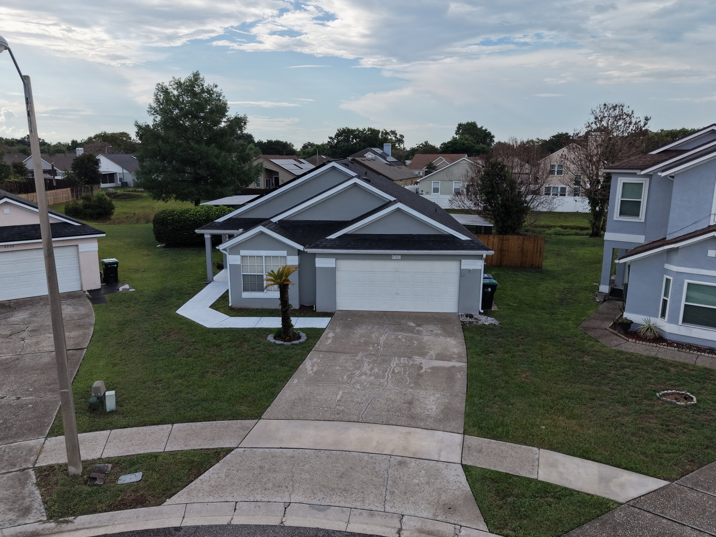 A suburban neighborhood with multiple houses, some with solar panels, a grassy yard, trees, a driveway, and a partly cloudy sky.