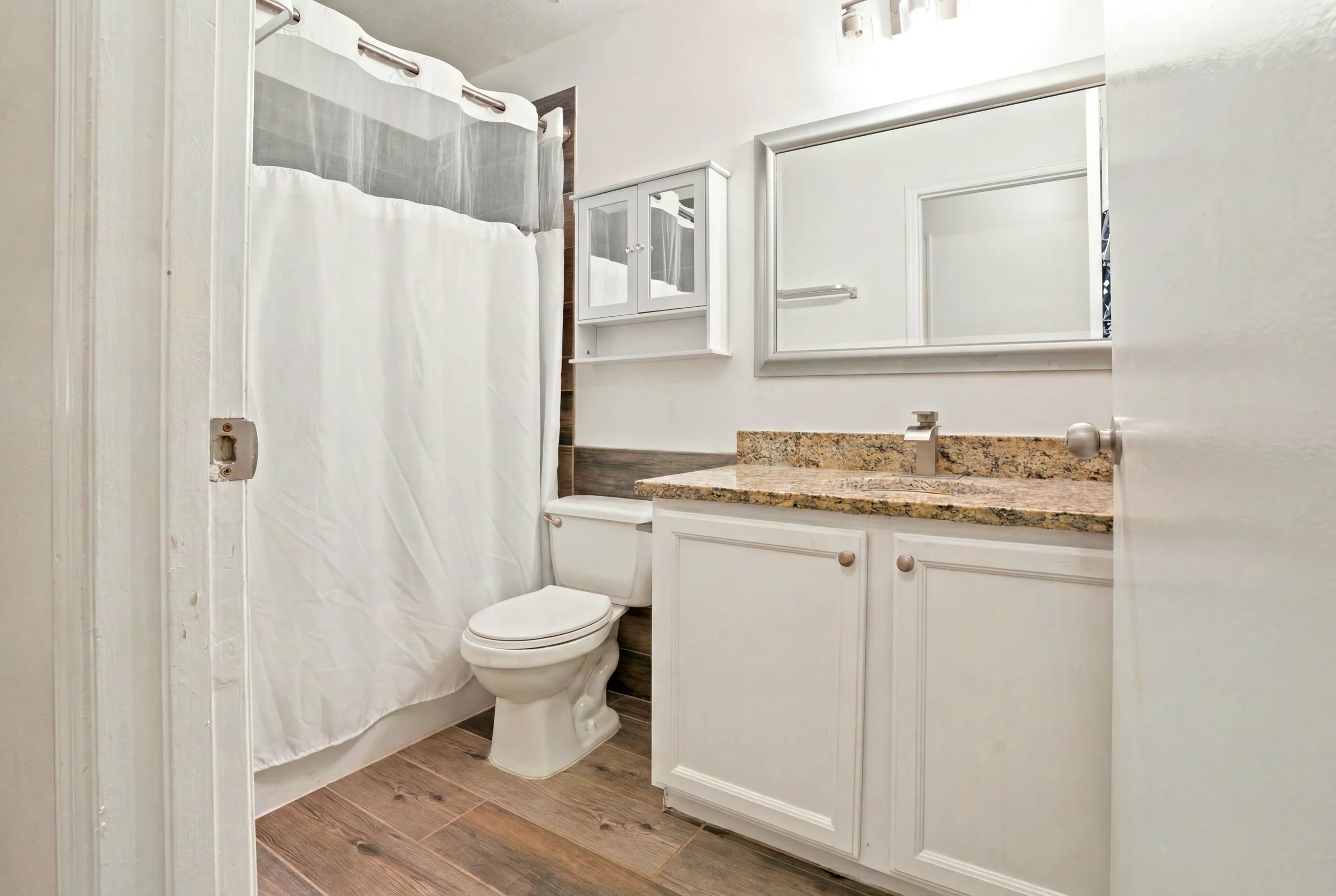Bathroom with a white vanity with granite countertop, mirror, toilet, shower with white curtain, and wall-mounted cabinet with mirrored doors.