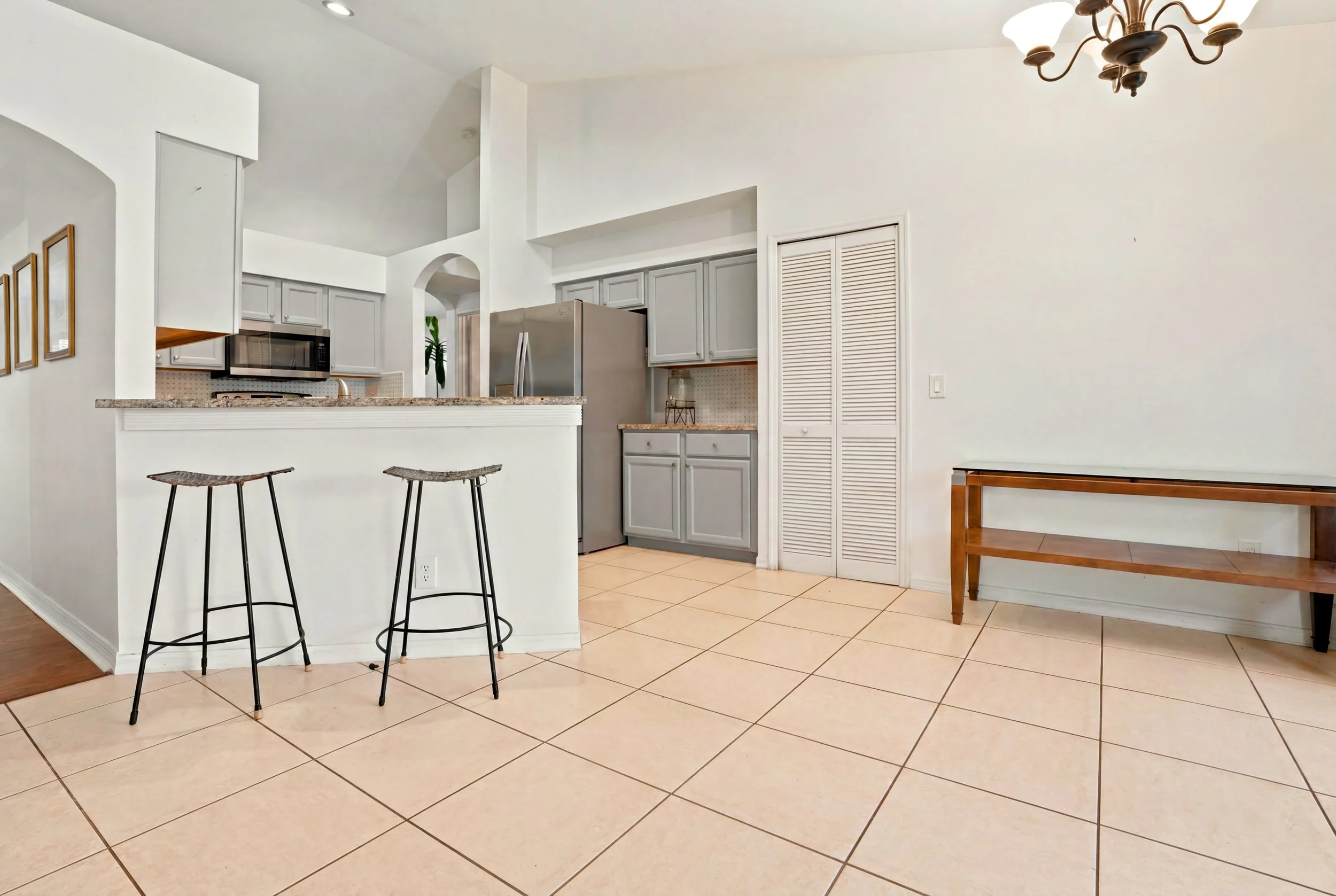 A kitchen with tiled floor, gray cabinets, a stainless steel refrigerator, a microwave, a small bar-style counter with two stool chairs, and a wooden table against a white wall with a chandelier overhead.