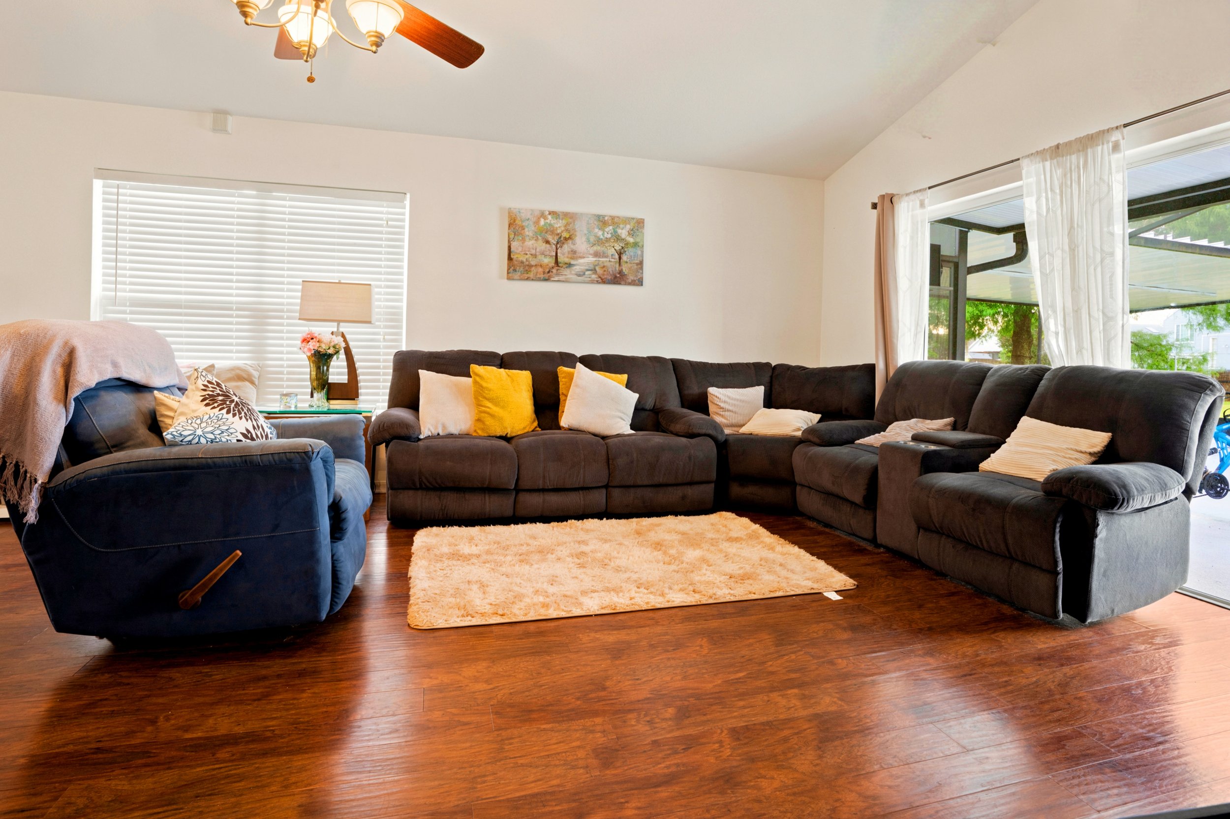 A living room with a large L-shaped dark sofa with white, yellow, and beige pillows, a small dark armchair with a blanket, a light pink rug, a wooden side table with a vase of pink flowers and a table lamp, white blinds on the window, a colorful land