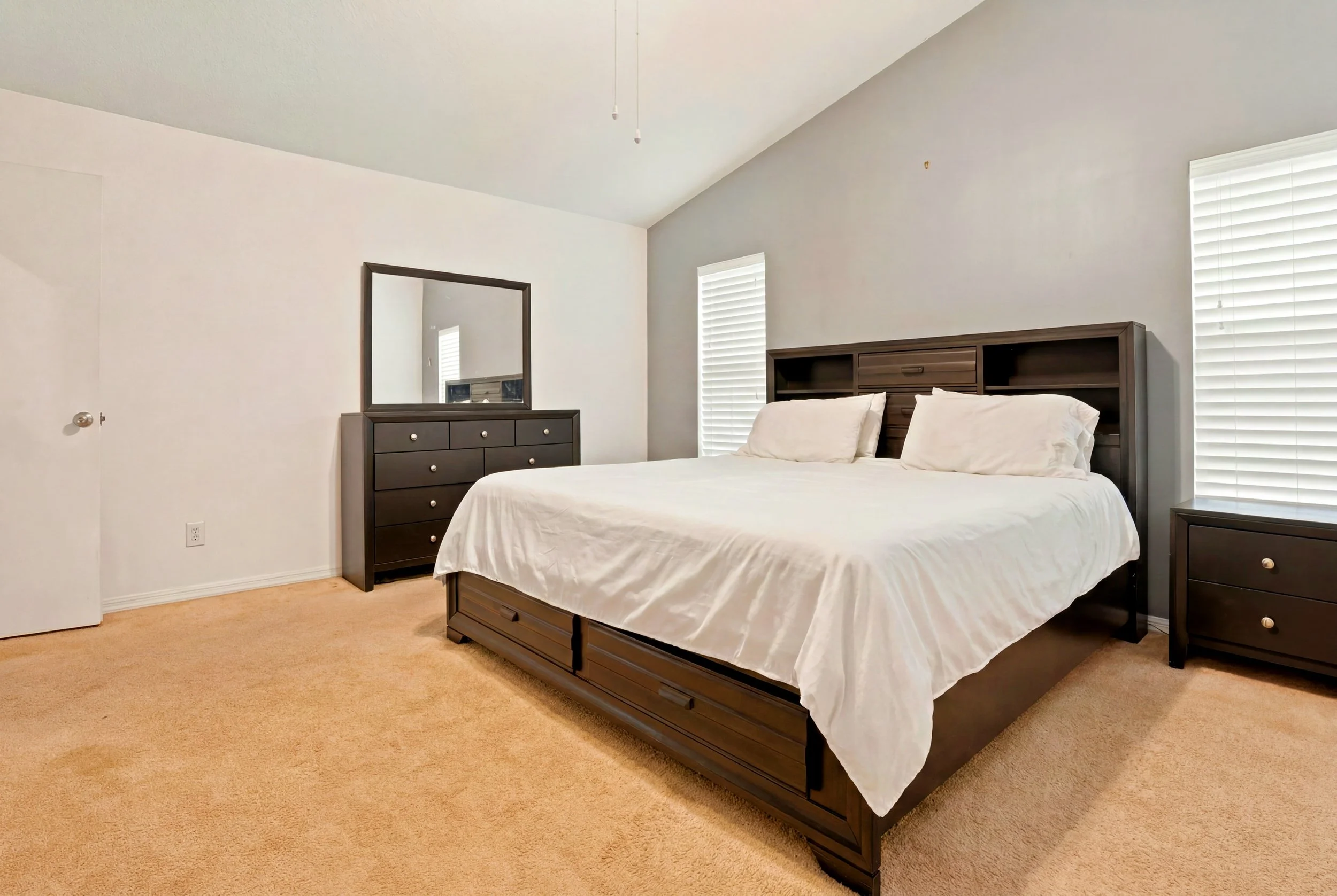 A bedroom with beige carpet, white walls, and a high sloped ceiling. There is a dark wooden bed with white bedding and pillows, a matching dresser with a mirror, and two windows with white blinds.