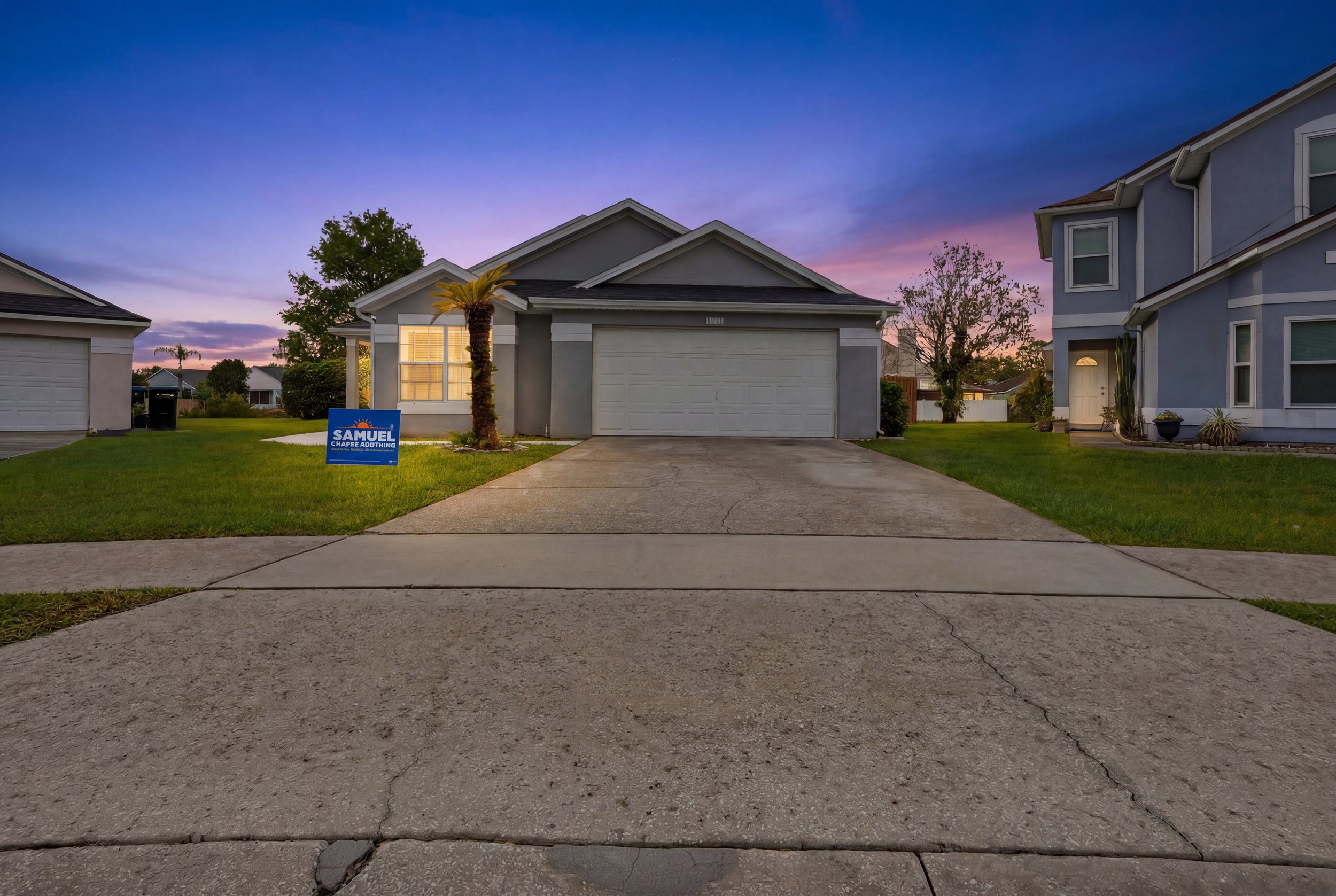 View of a suburban house at dusk with a purple and pink sky, a palm tree, and a campaign sign in the front yard. The house has a closed garage door and a well-maintained lawn.