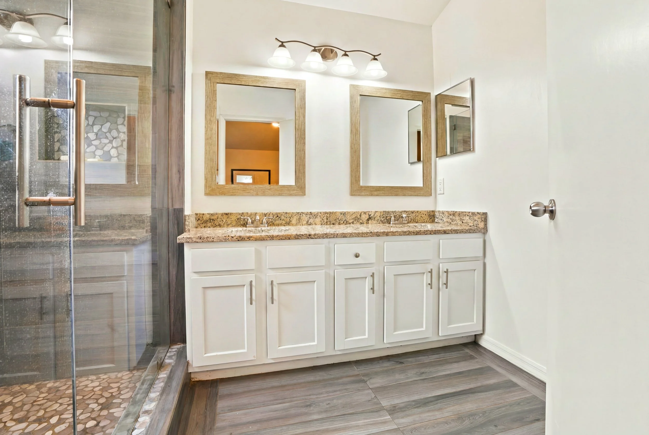 Bathroom with a double sink vanity with white cabinets, a granite countertop, and two mirrors above. To the left, there is a glass shower door with a towel bar and pebble stone shower floor. The wall is painted white, and there is a light fixture wit