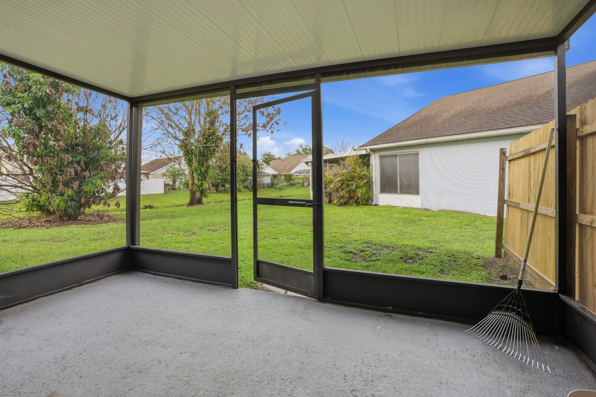 View from a screened porch looking out onto a backyard with green grass, trees, and neighboring houses under a blue sky.