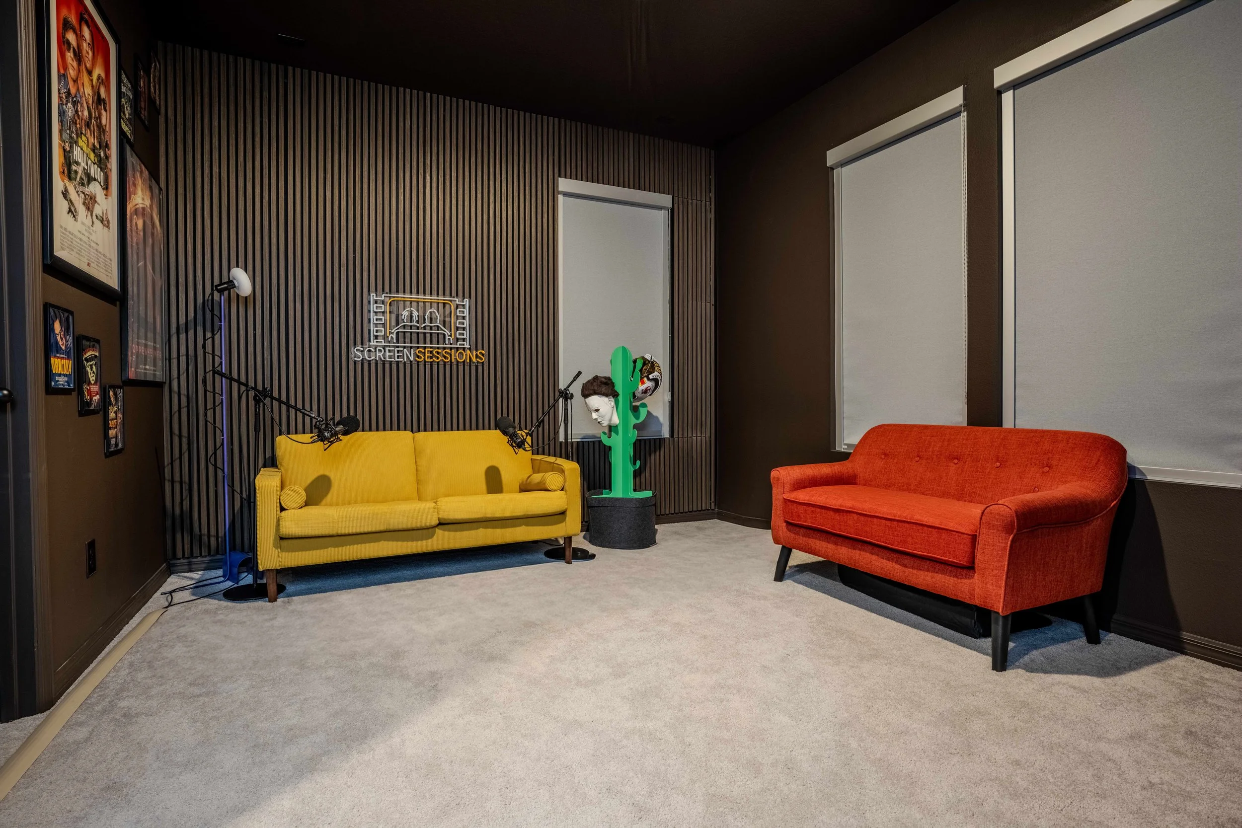 A cozy room with two colorful sofas, a yellow one and an orange one. The wall has movie posters and a neon sign that reads 'SCREEN SESSIONS.' There are window blinds and a cactus-shaped coat rack with a decorative mask hanging on it. The floor is car