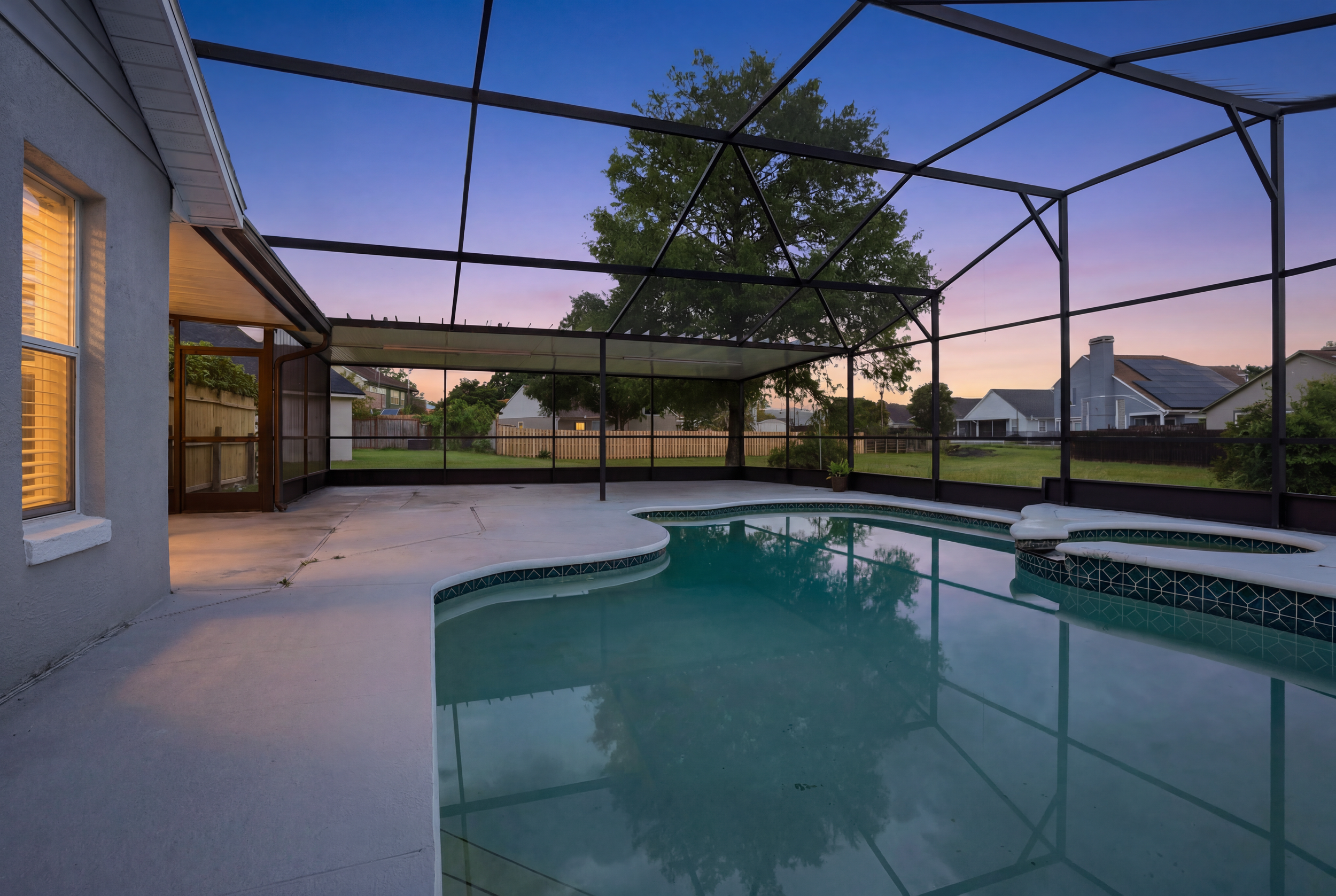 A backyard pool area at dusk with a screened enclosure, a large tree, and neighboring houses visible in the background.