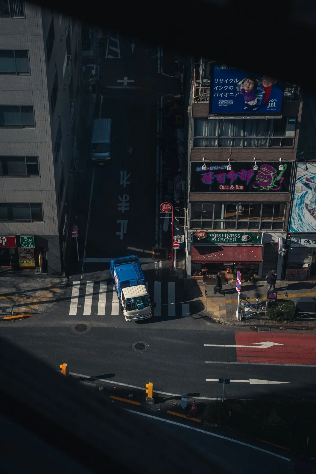 View from a tall building showing a city street with parked cars, pedestrians, and buildings with colorful signs, including a burger shop, a nightclub, and a billboard with two cartoon characters in purple and red clothing.