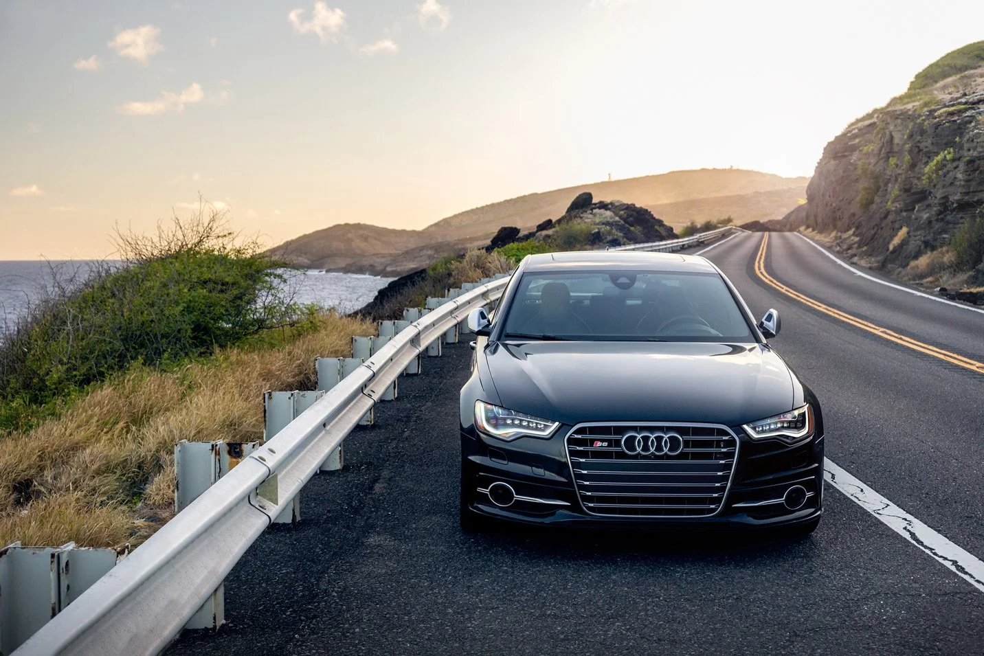 A black Audi car parked on the side of a scenic coastal road with a guardrail, overlooking the ocean and rolling hills at sunset.