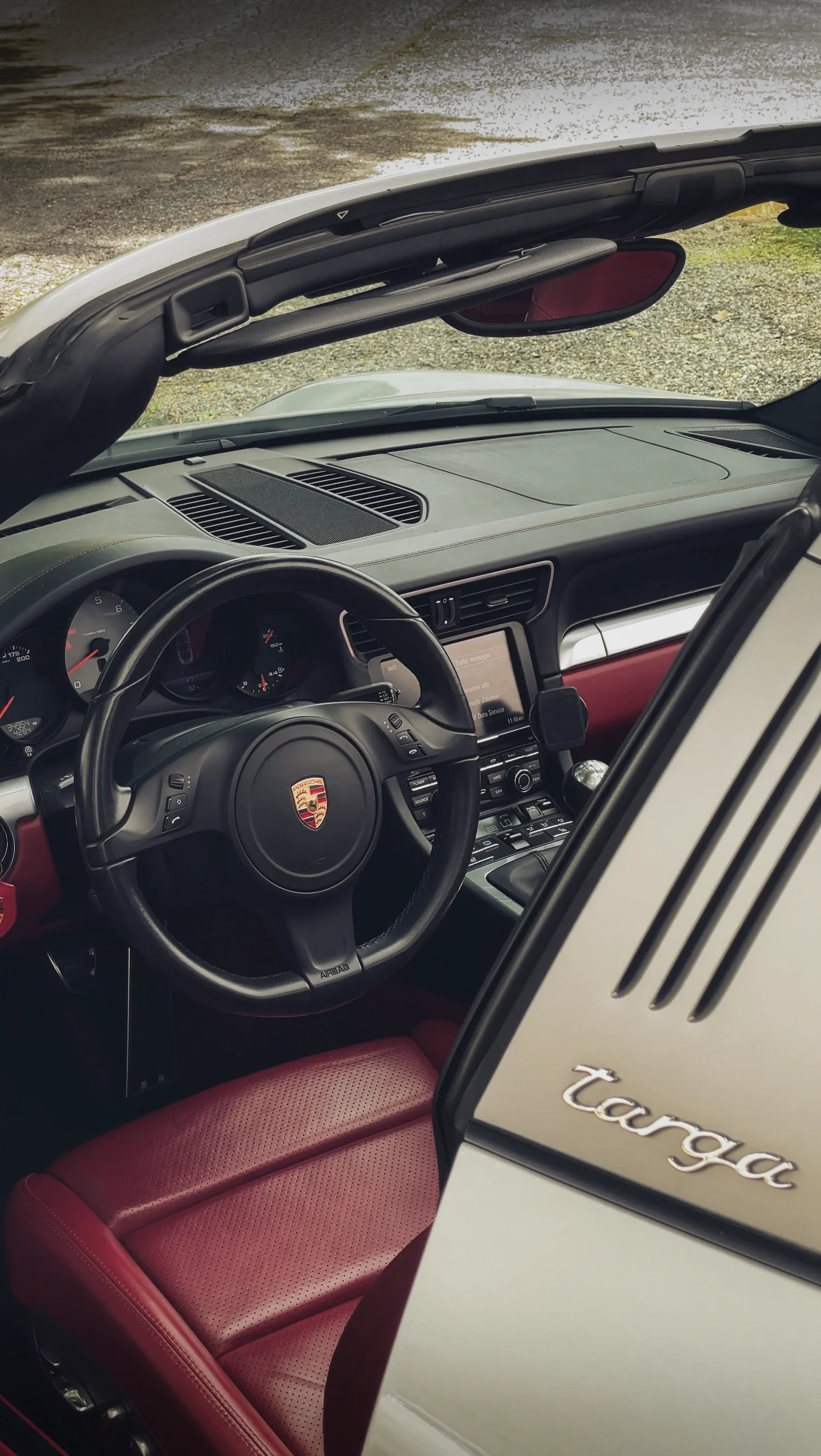 Interior of a Porsche 911 Targa with a black steering wheel, dashboard, digital display, and red seating, with a partial view of the open roof and a Porsche Targa emblem.