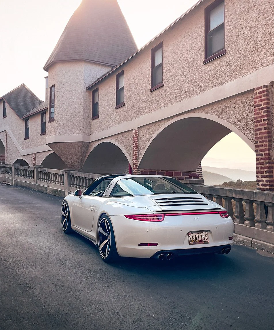 A white Porsche 911 parked on a street under an arched building with a turret, with a scenic view in the background.