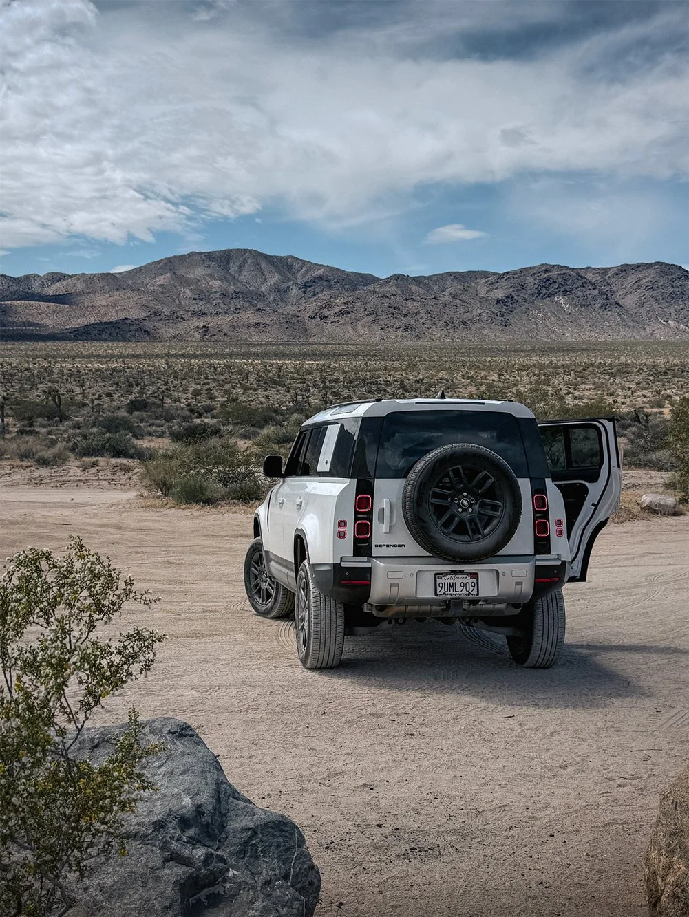 An off-road white Land Rover Defender parked on a dirt path in a desert landscape with mountains in the background and a partly cloudy sky.