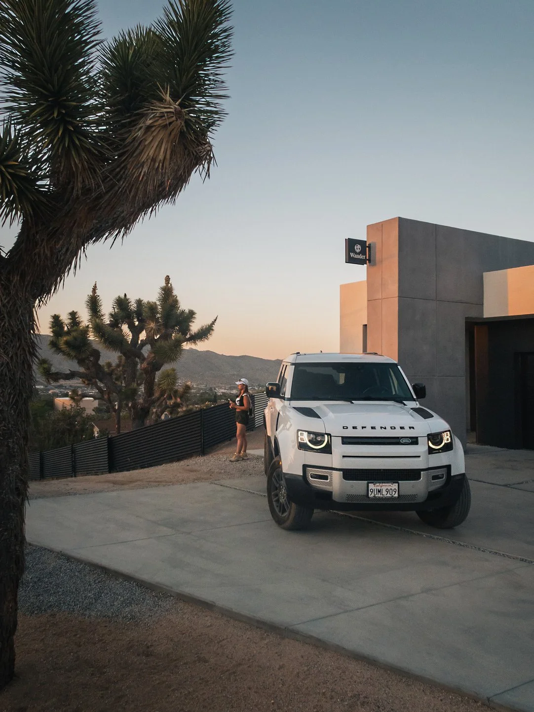 A white Land Rover Defender parked outside a modern building with a person standing nearby, in a desert landscape with large cacti and mountains in the background during sunset.