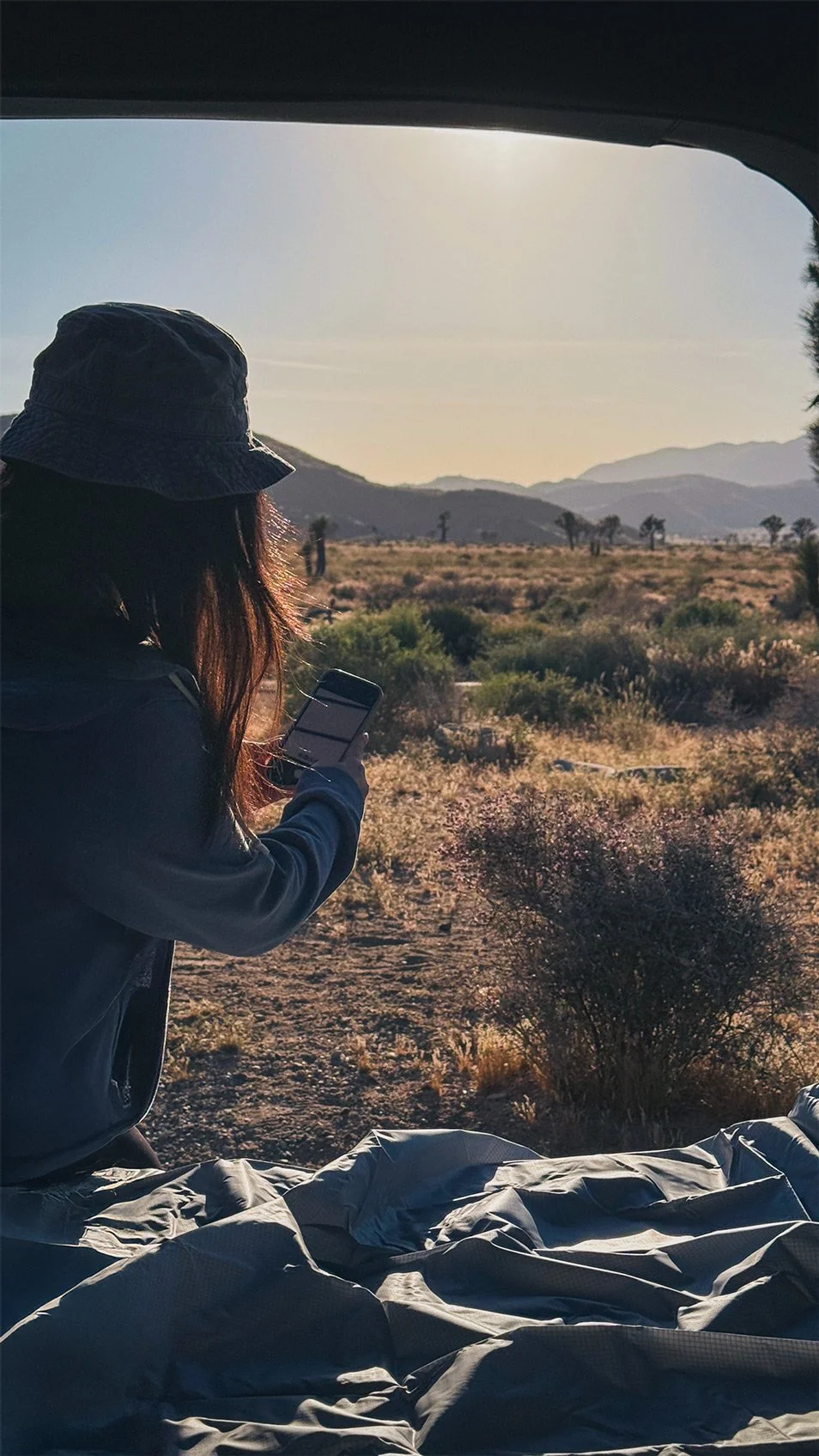 Person wearing a hat and jacket sitting on a sleeping bag in a desert landscape, looking at their phone, with mountains and sparse vegetation in the background under a clear sky.