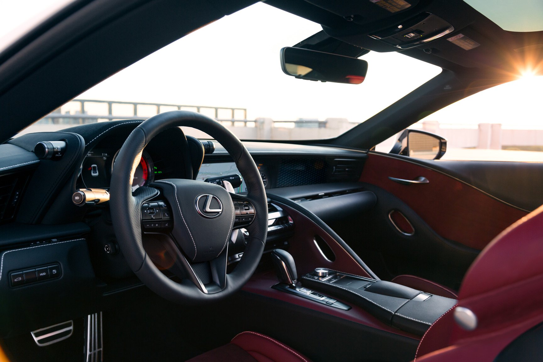 Interior of a Lexus LC500 showing the steering wheel, dashboard, and center console with gear shifter, with a view of the road and sunset through the windshield.