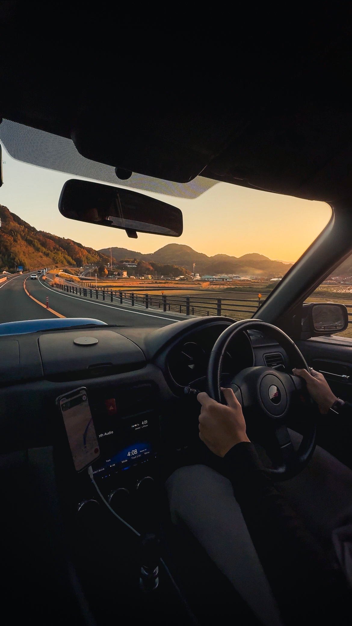 Perspective from inside a car showing the driver’s hands on the steering wheel, with a scenic road and mountains in Japan outside during sunset.