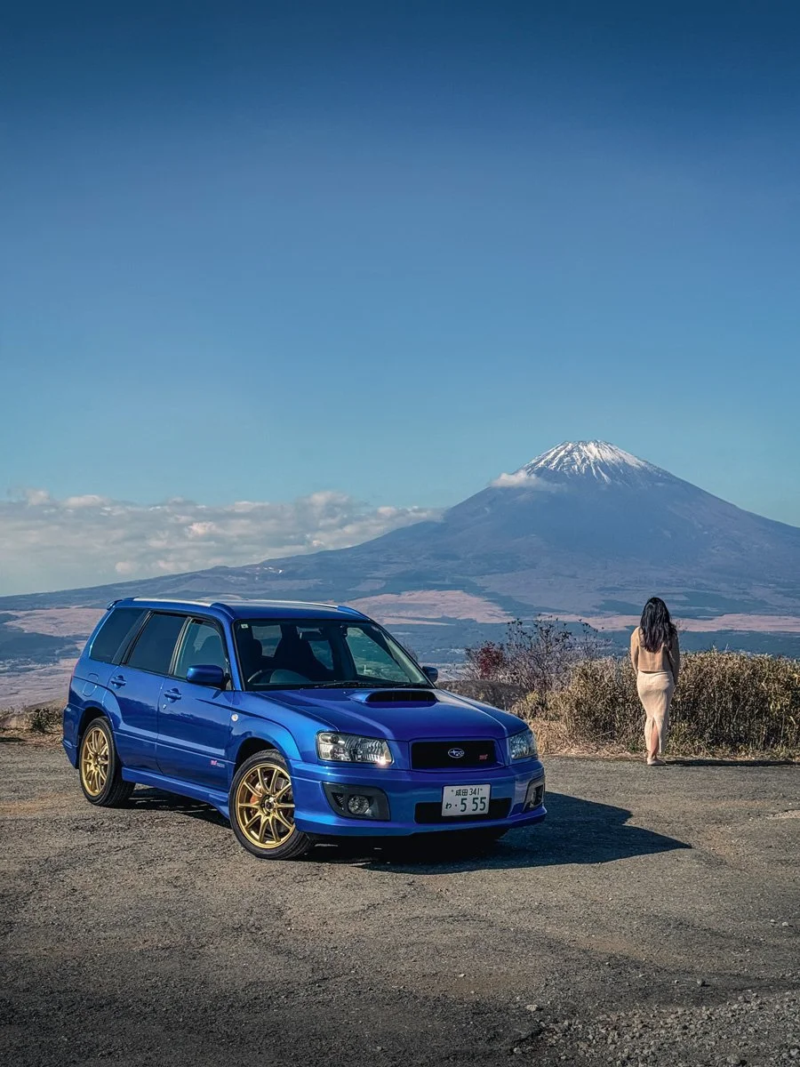 A blue Subaru Forester STi with gold wheels parked on a dirt road with Mount Fuji and a woman in a beige dress walking away in the background.