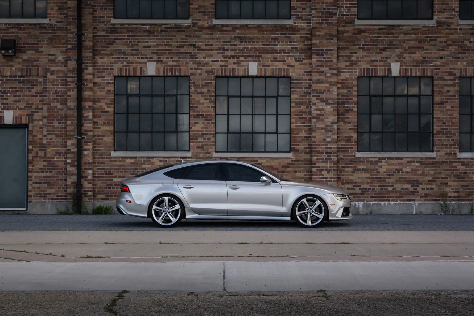 Silver luxury sedan car parked beside a brick industrial building with large grid-style windows.