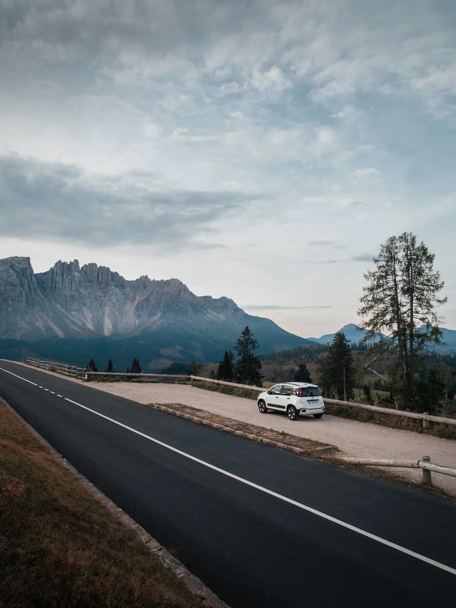 A scenic mountain landscape with a winding black asphalt road in the foreground, a white SUV parked on a gravel turnout, tall evergreen trees, and rugged mountain peaks under a partly cloudy sky.