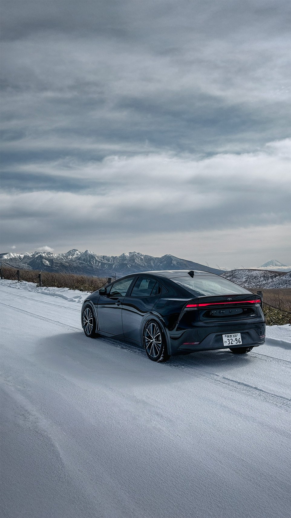 Black electric car parked on a snowy mountain road with snow-capped mountains in the background and cloudy sky overhead.
