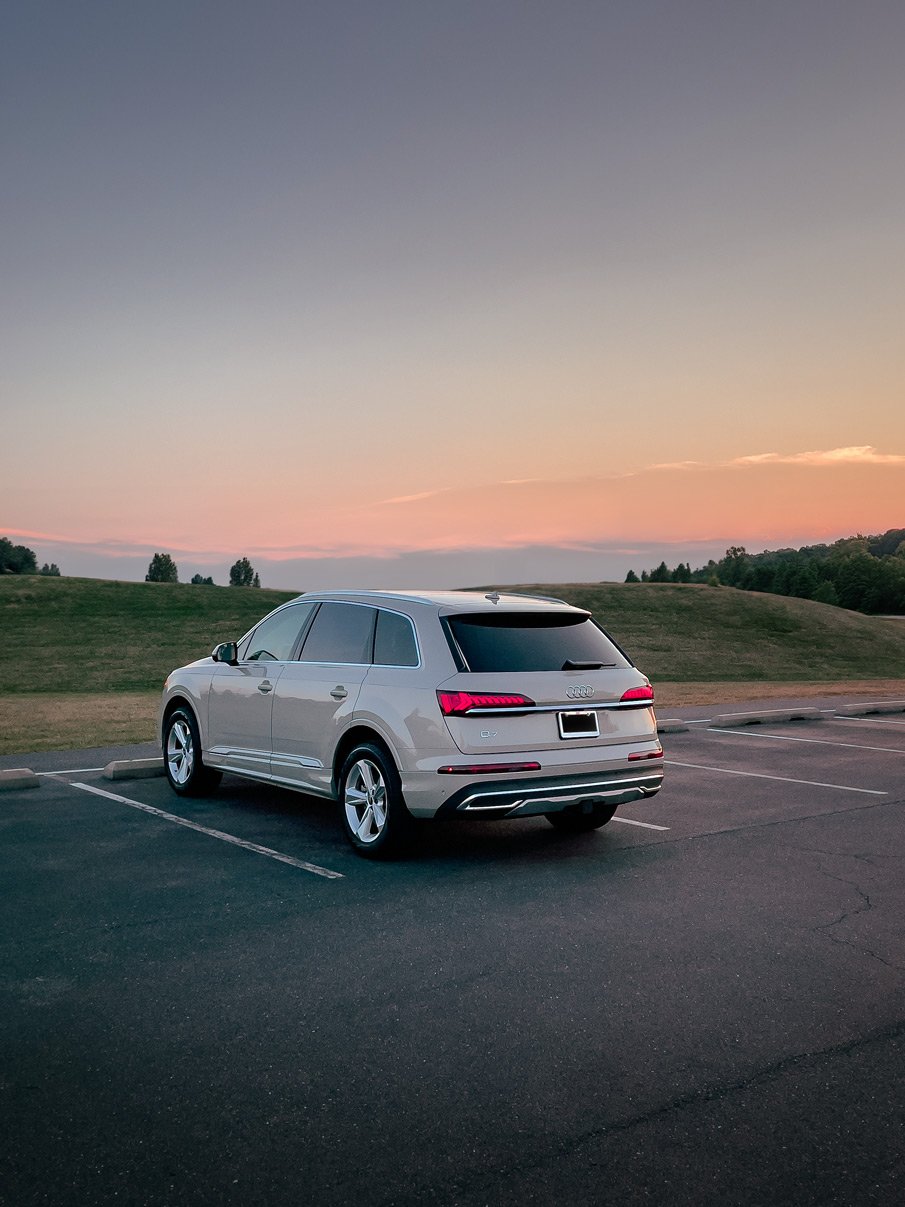 Silver Audi Q7 parked in an empty parking lot during sunset with grassy hills and a pink and purple sky in the background.