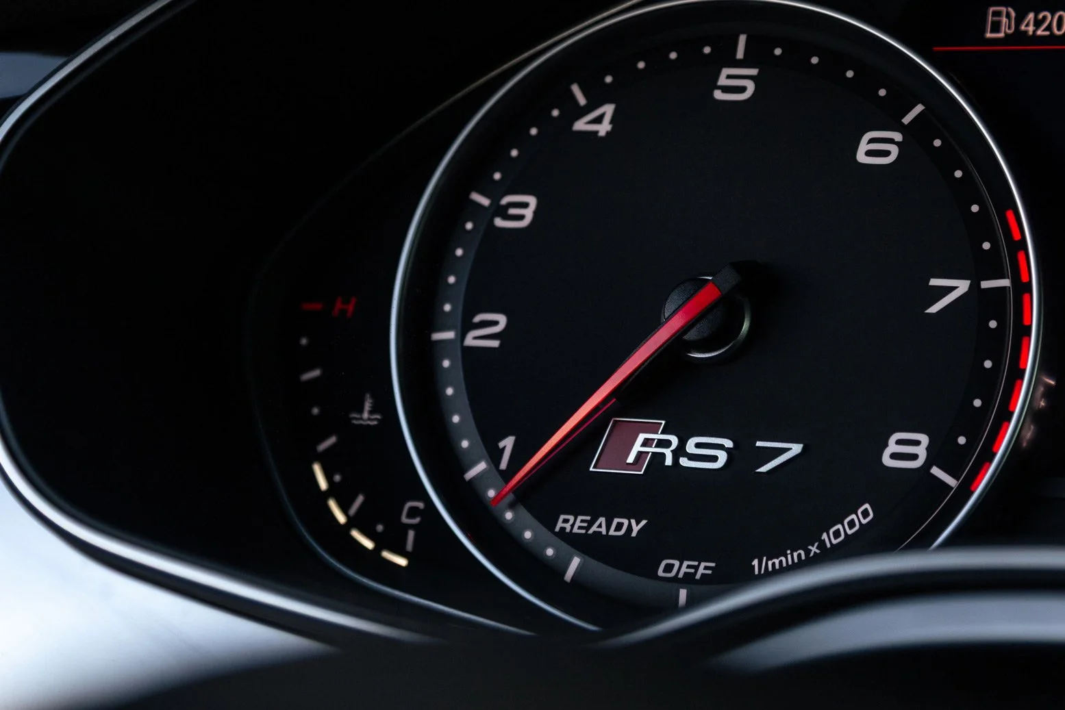 Close-up of a car dashboard showing a tachometer with the needle pointing to 1, and a small temperature gauge on the left, with a red and white 