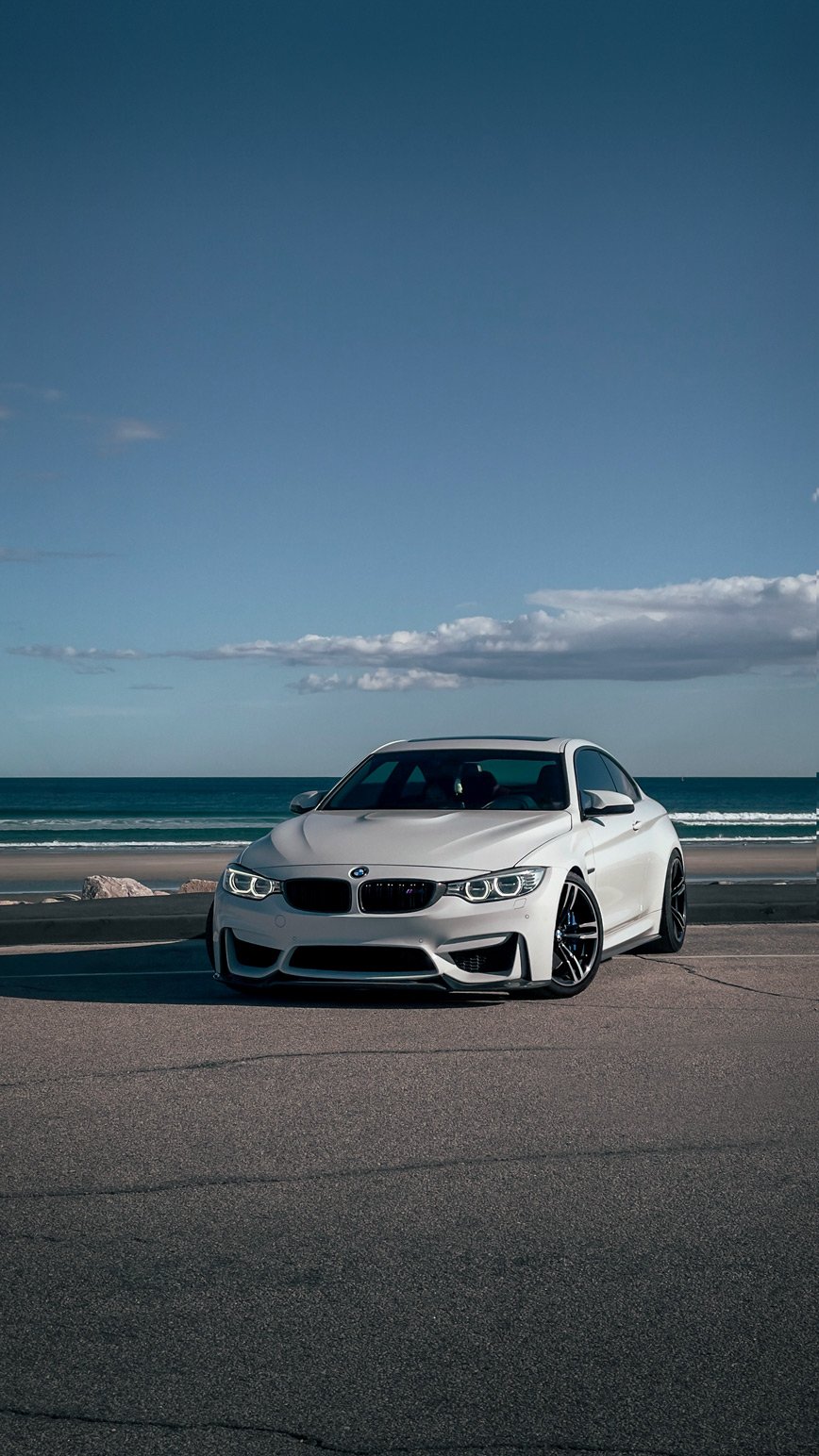 A white BMW M4 coupe parked on the street near a beach with the ocean and blue sky with some clouds in the background.