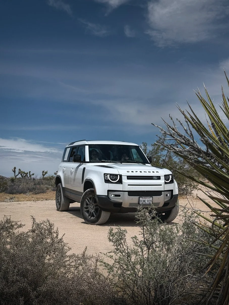 A white Land Rover Defender parked on a sandy dirt road in a desert landscape with sparse desert plants and a blue sky with wispy clouds.