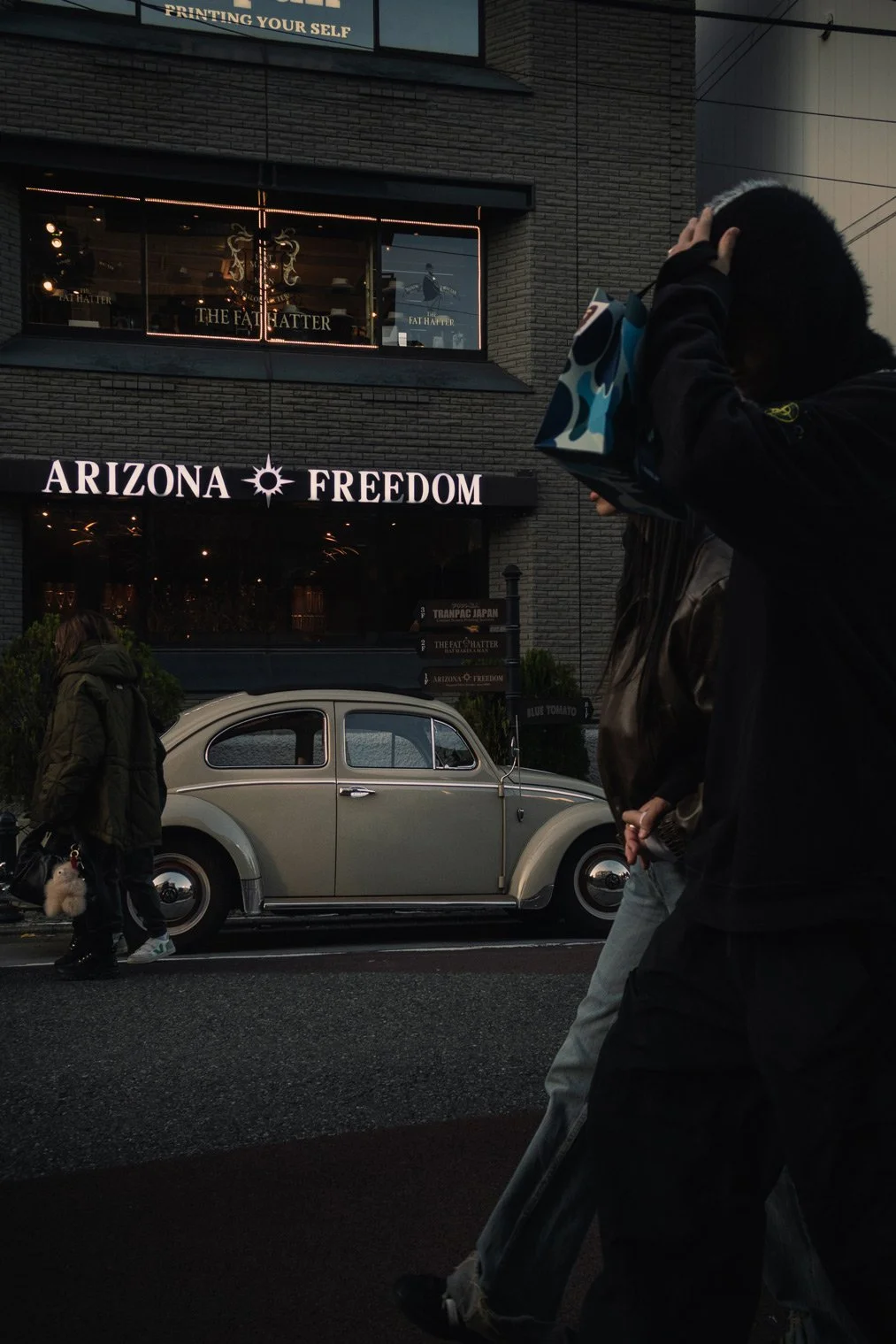 Street scene with vintage Volkswagen Beetle parked in front of a building with illuminated signs that read 'ARIZONA * FREEDOM' and 'THE FADHATTER.' Several pedestrians walk along the sidewalk, one person on the right side holding a shopping bag and a