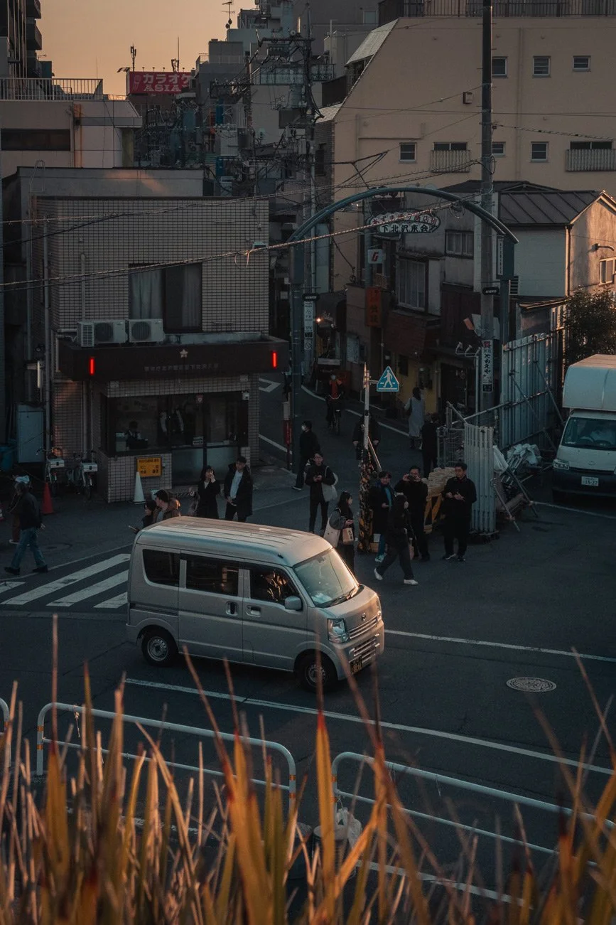 An urban street scene during dusk in Japan, with pedestrians crossing at a crosswalk, a silver van in the foreground, and various buildings, power lines, and signage in the background.